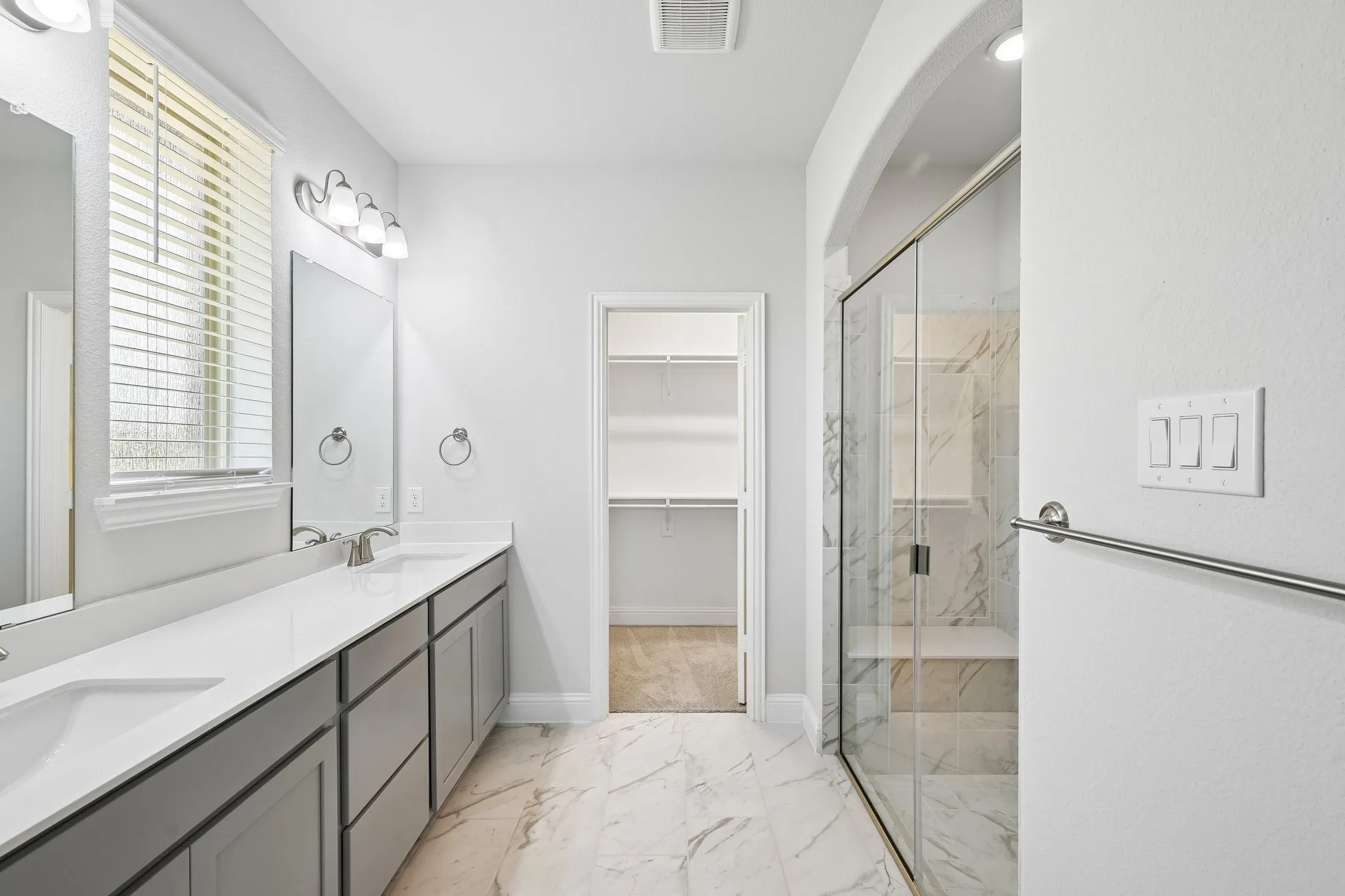 Bathroom featuring a double vanity with gray cabinetry, a walk-in shower with clear glass enclosure and stone-look tile, and a spacious walk-in closet
