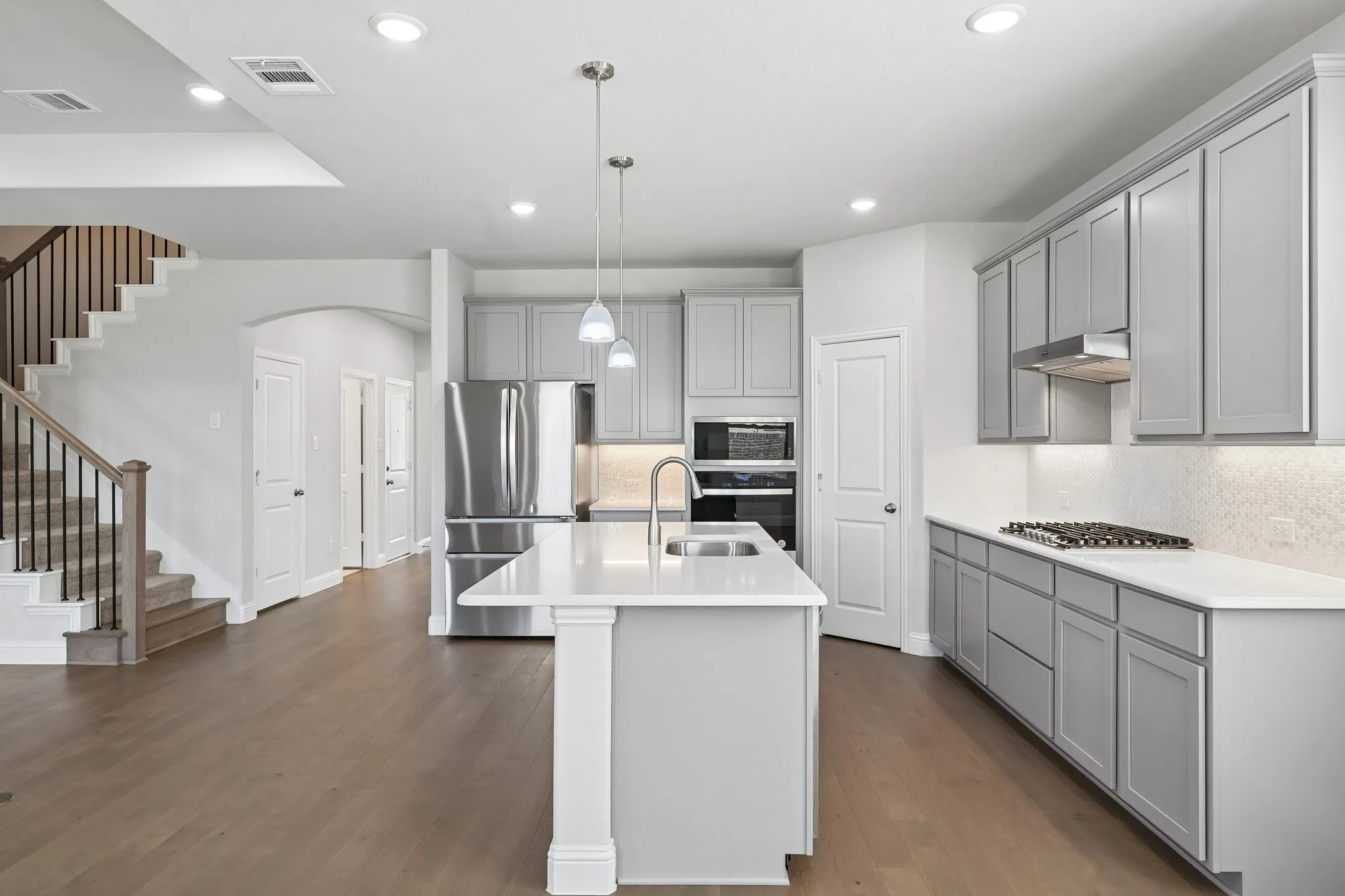 Open-concept kitchen featuring an island with sink and pendant lighting, light gray cabinetry, stainless steel appliances, and wood-finish flooring