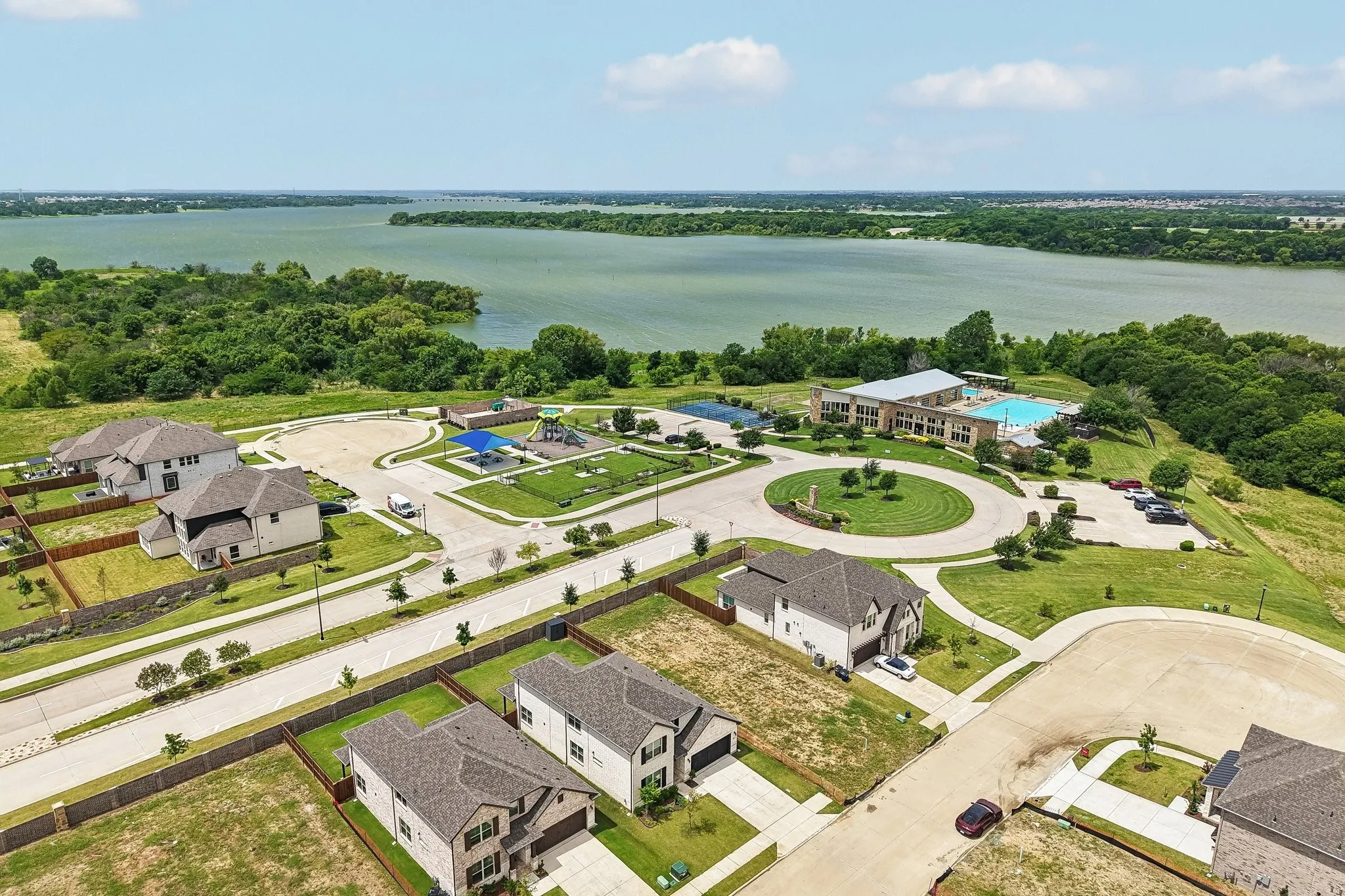 Aerial perspective showcasing residential properties, a central community park with recreational equipment, a community building with an outdoor pool, and a large body of water bordered by extensive greenery