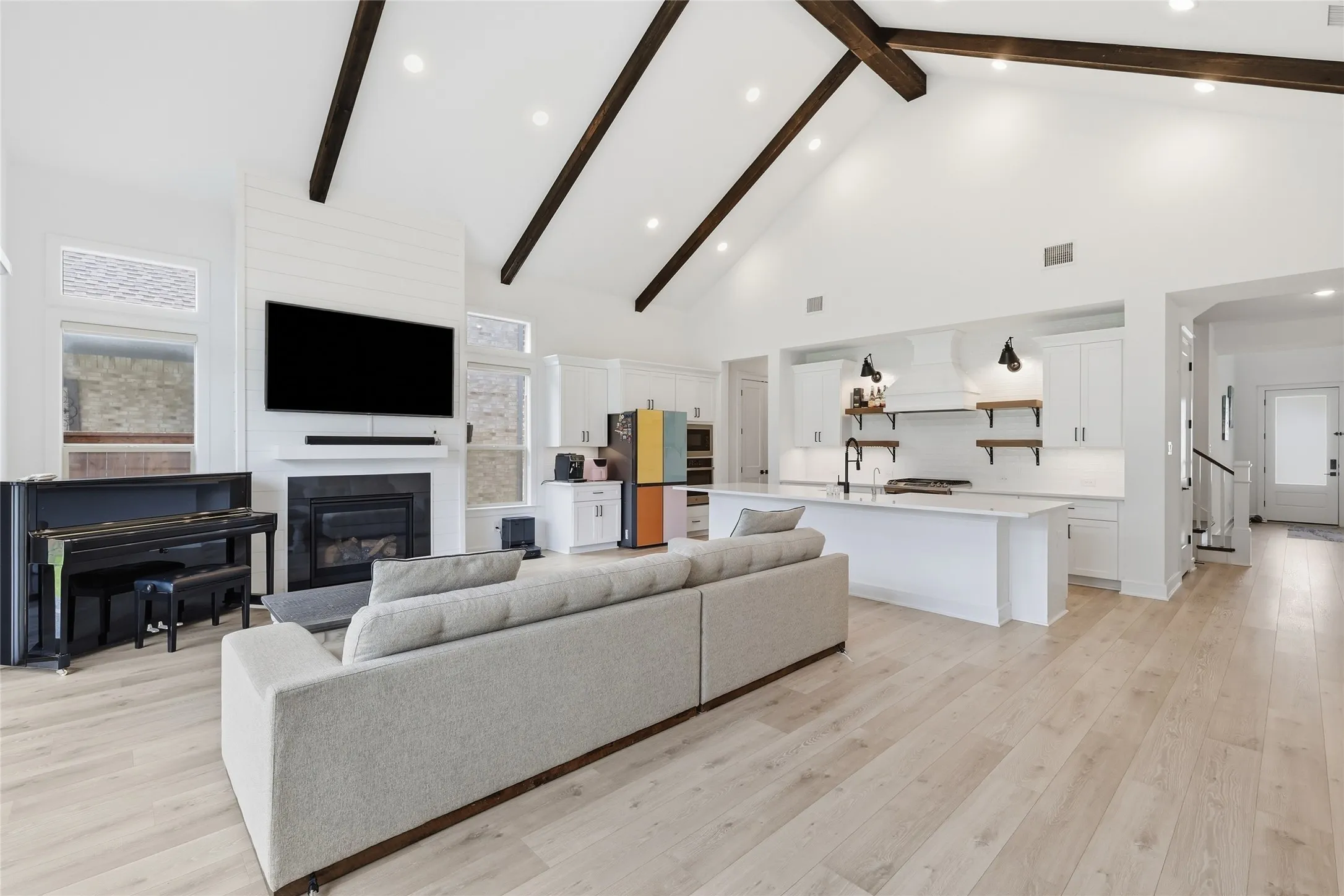 Open-concept living area featuring a vaulted ceiling with exposed wood beams, light wood-finish flooring, and a fireplace with a shiplap accent wall