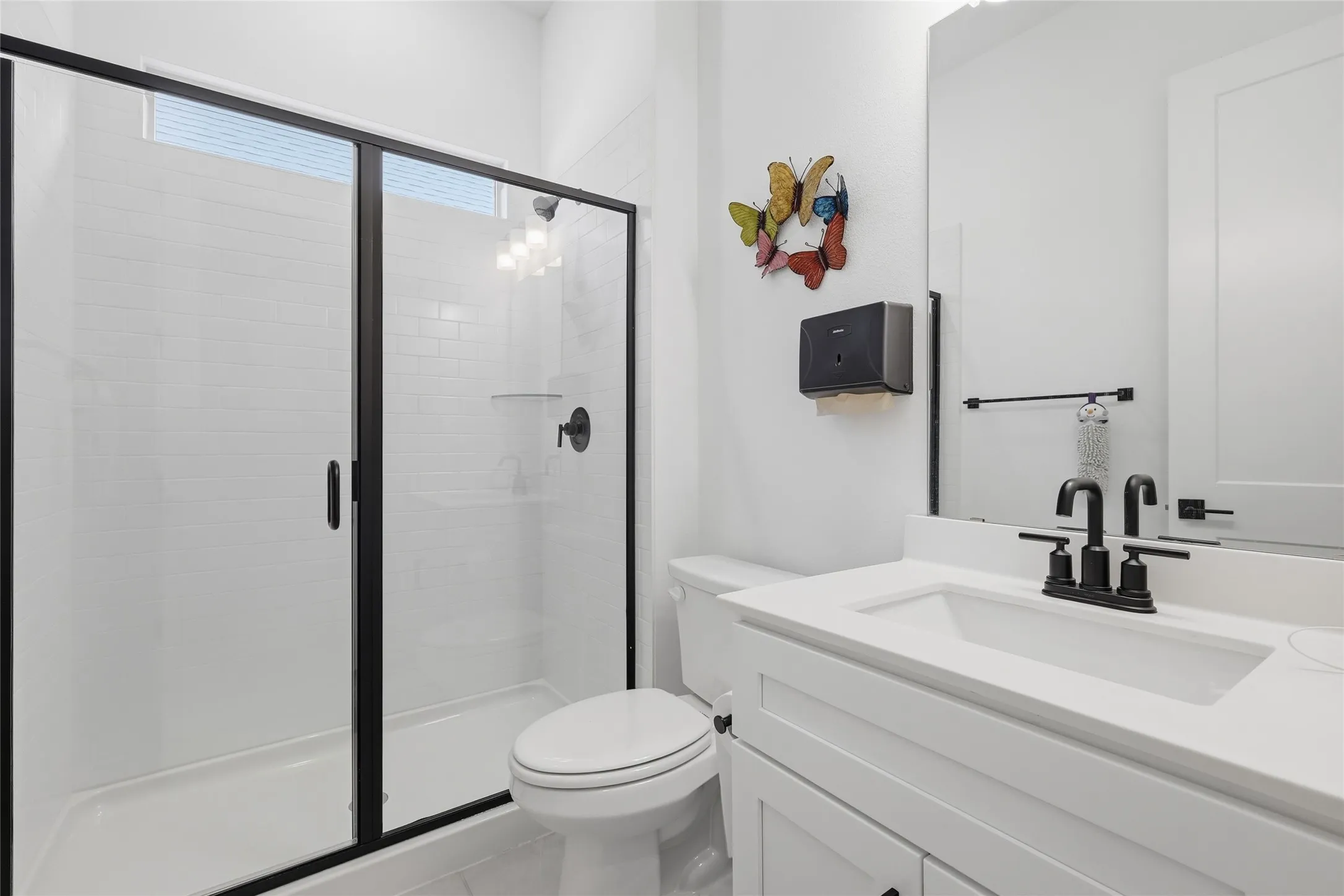 Modern bathroom featuring a walk-in shower with black-framed glass enclosure and white subway tile surround, a single vanity with white countertop and integrated sink, matte black faucet, and a large wall-mounted mirror