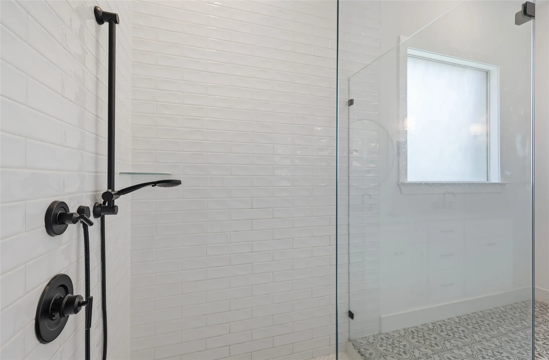 Walk-in shower featuring matte black fixtures, white subway tile surround, clear glass enclosure, and patterned floor tile