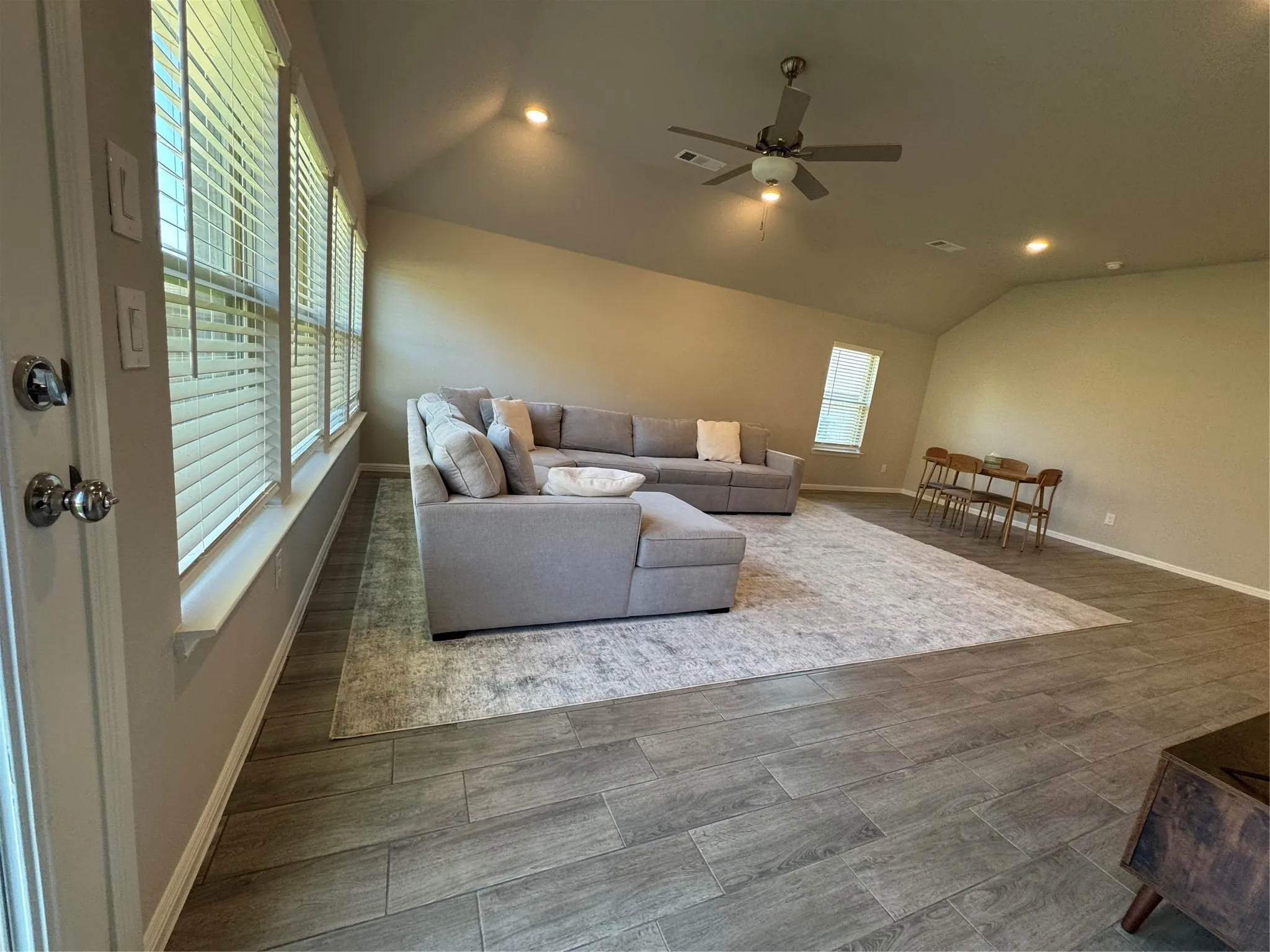 Open-concept living space with wood-finish tile flooring, vaulted ceilings, recessed lighting, and multiple windows with blinds