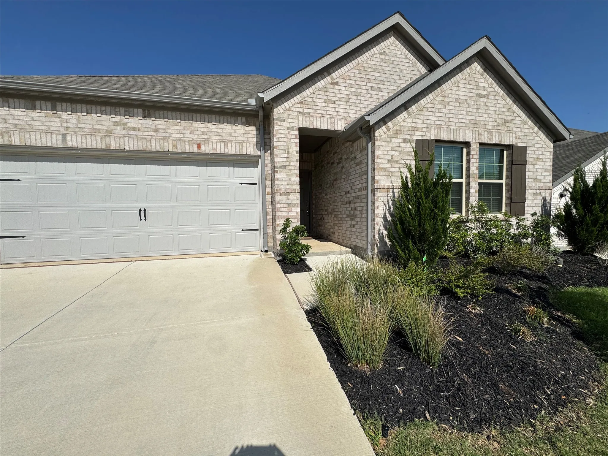 Light-colored brick exterior with a two-car garage, dark decorative window shutters, and low-maintenance landscaping with dark mulch