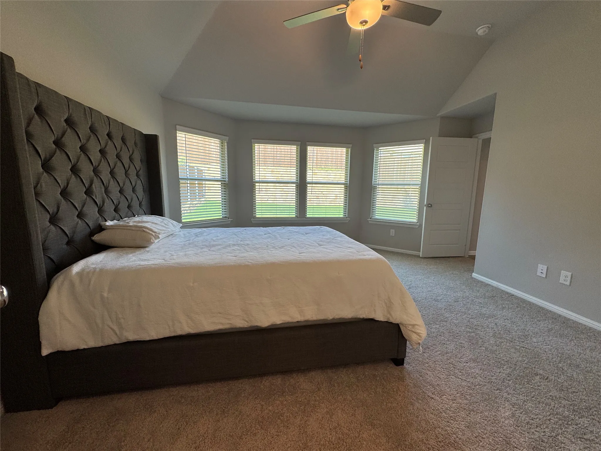 Spacious room featuring a vaulted ceiling with a ceiling fan, a bay window with blinds, and soft carpet flooring