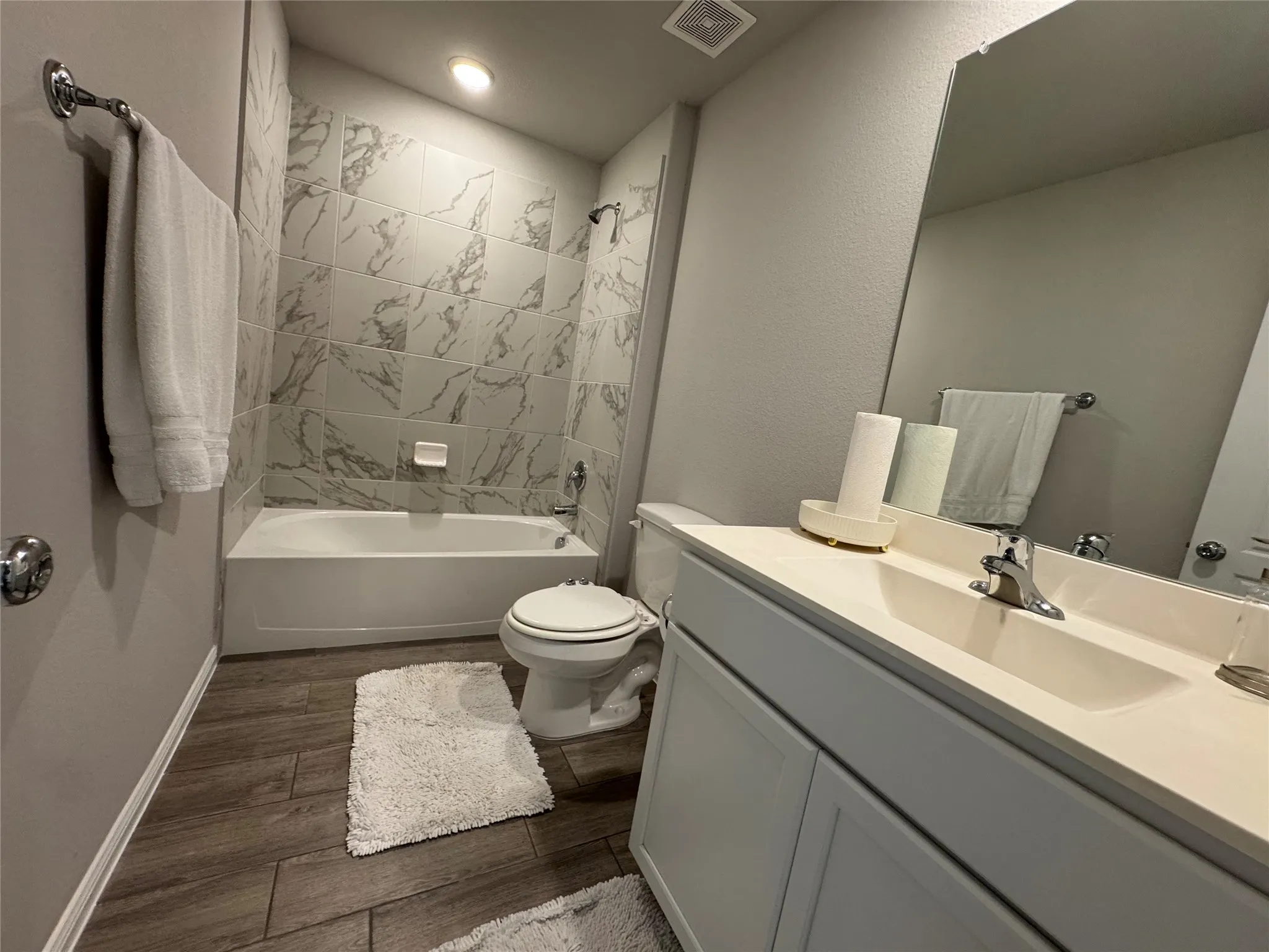 Bathroom featuring a built-in bathtub with a tiled surround, a white vanity with an integrated sink, a large wall mirror, and wood-finish flooring