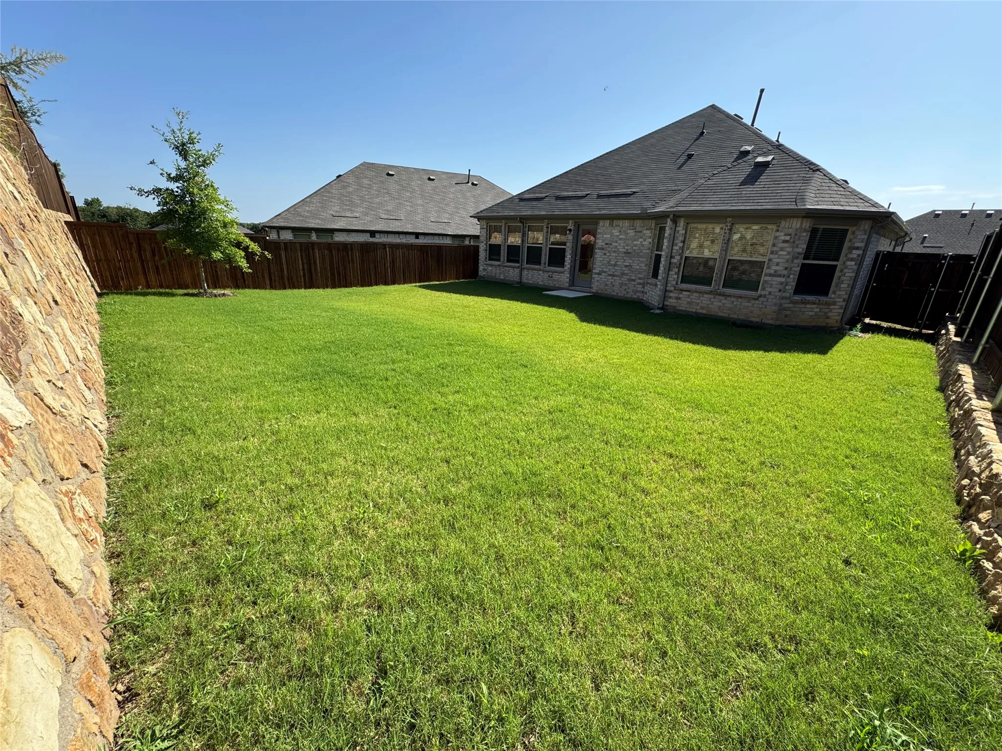 Expansive backyard featuring a lush green lawn, full privacy fencing, and stone retaining wall