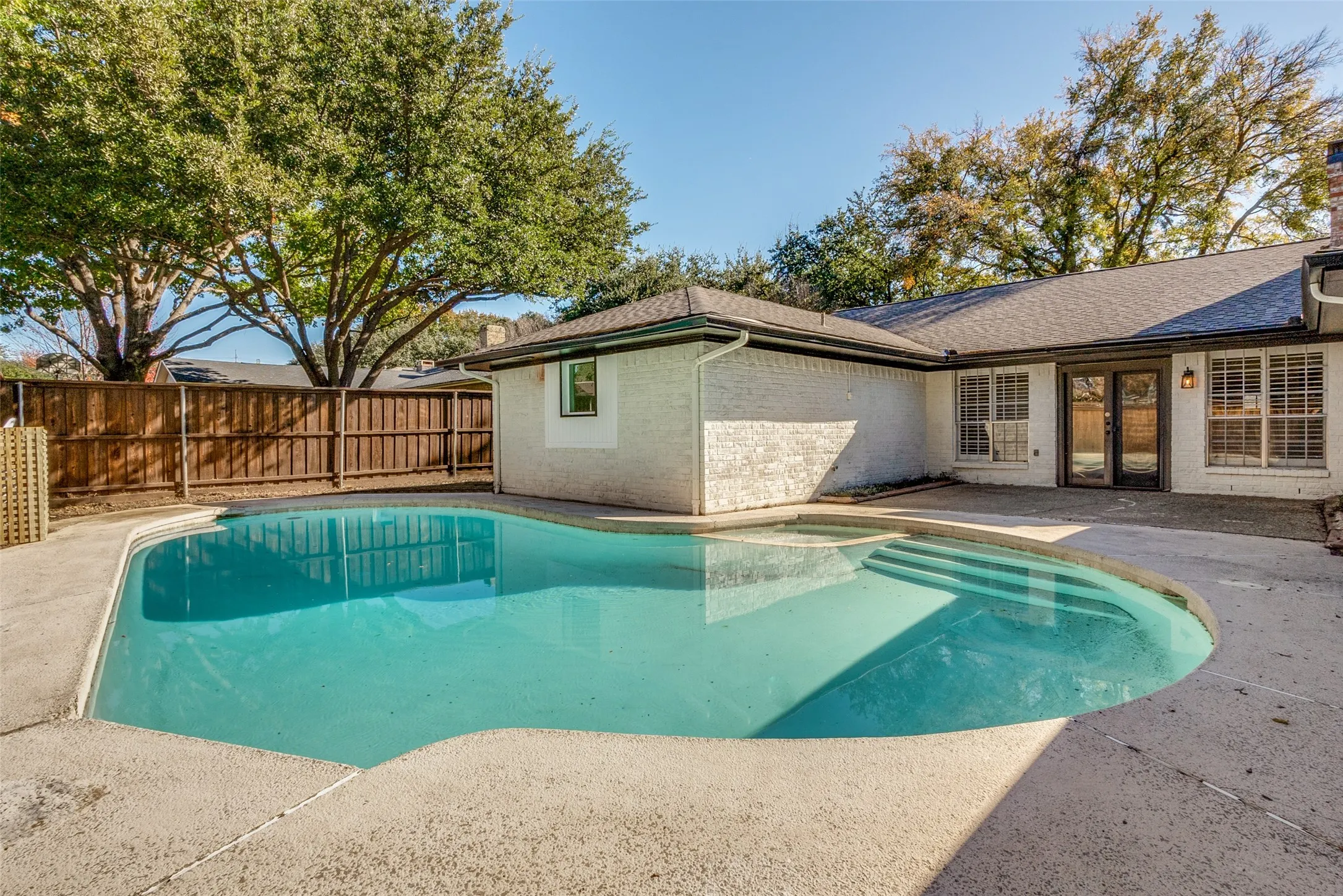 Inviting backyard featuring a freeform swimming pool with integrated steps, concrete decking, a privacy fence, and mature trees