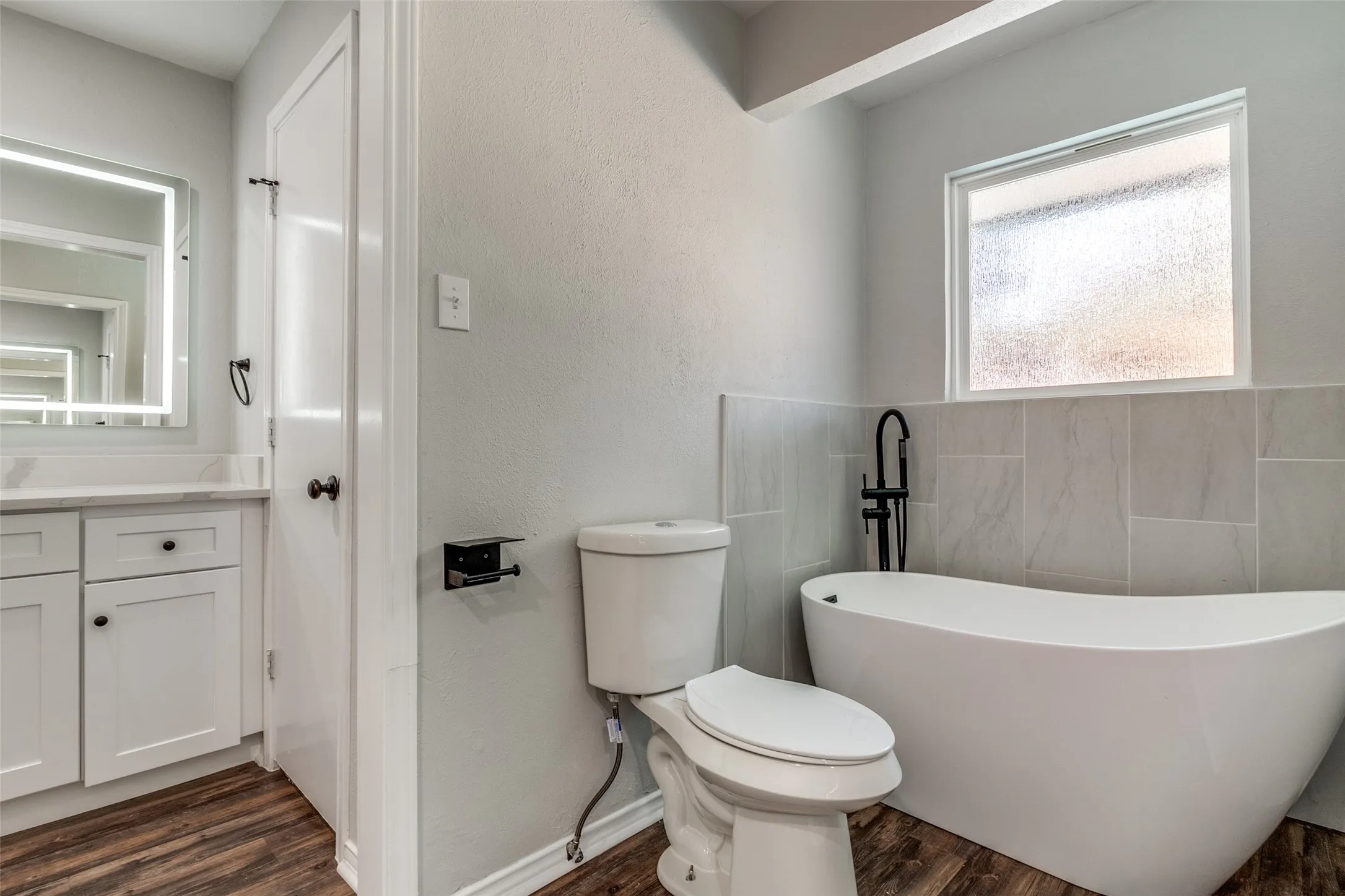 Freestanding soaking tub with matte black floor-mounted faucet, complemented by a white vanity with quartz countertop and illuminated mirror
