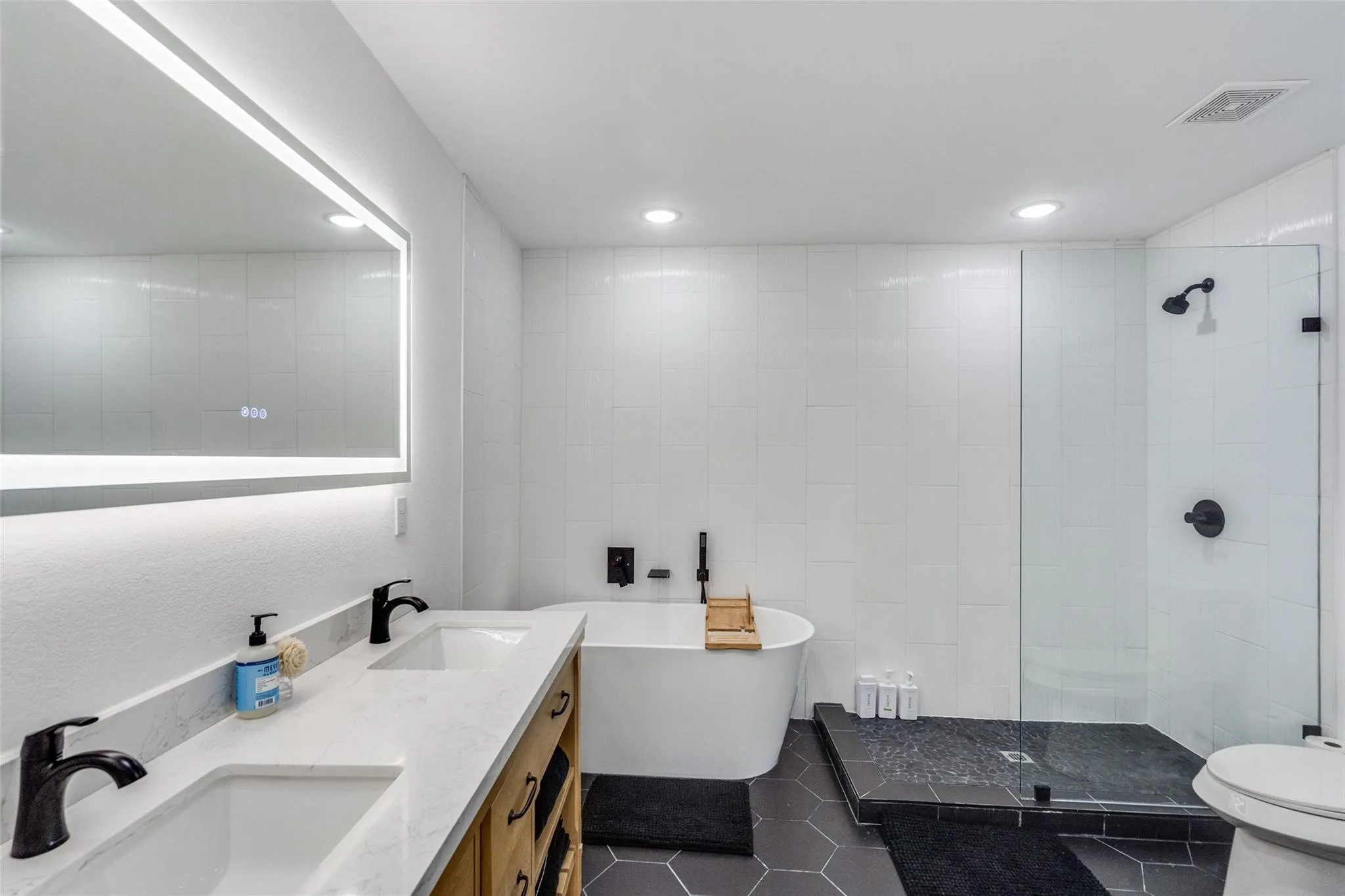 Contemporary bathroom featuring a dual vanity with light-colored countertops, a freestanding soaking tub, a glass-enclosed shower with dark fixtures, hexagon tile flooring, and an illuminated vanity mirror