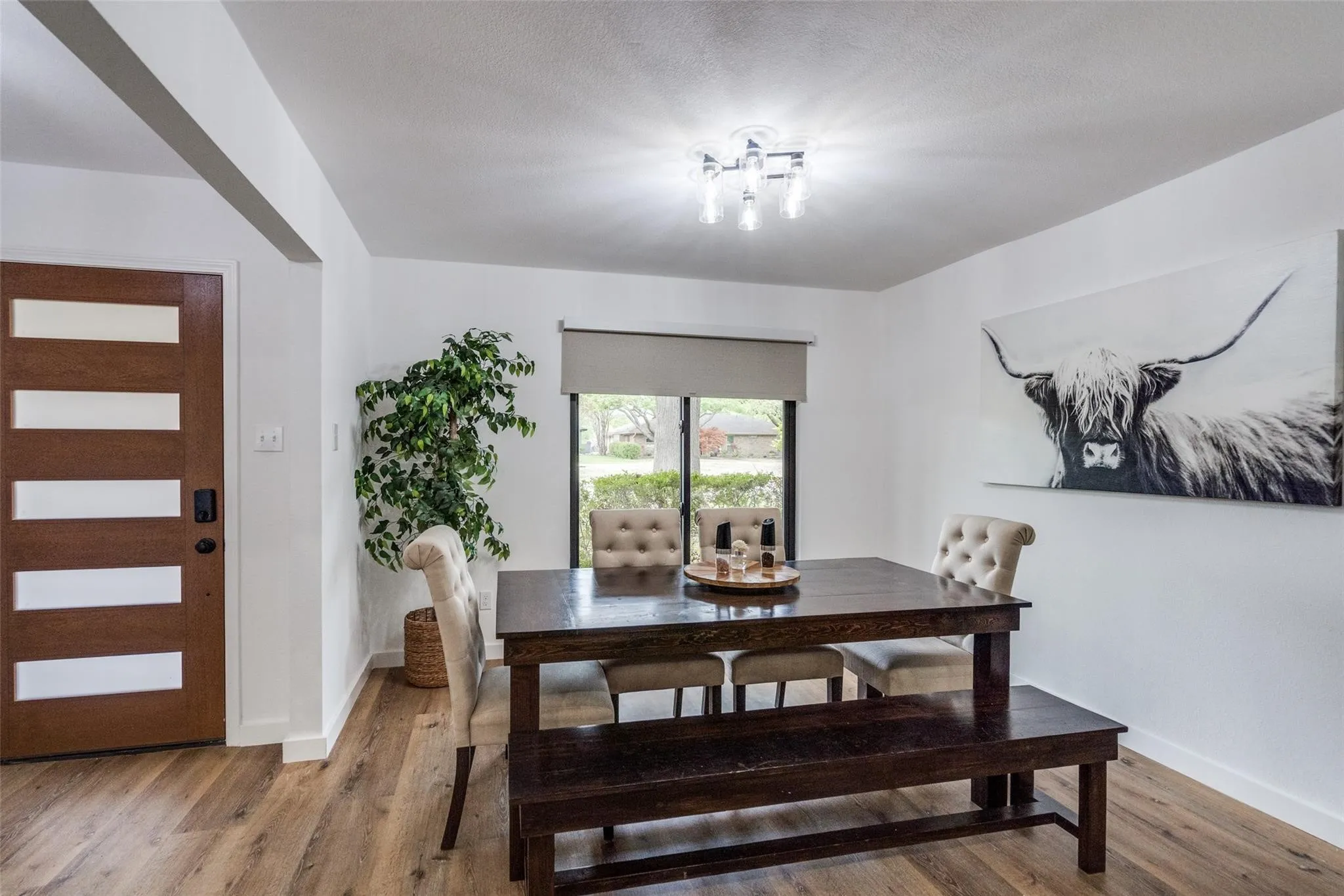 Inviting dining space featuring wood-finish flooring, a modern multi-bulb ceiling fixture, a window with a roller blind, and a wood-paneled door with horizontal frosted glass inserts