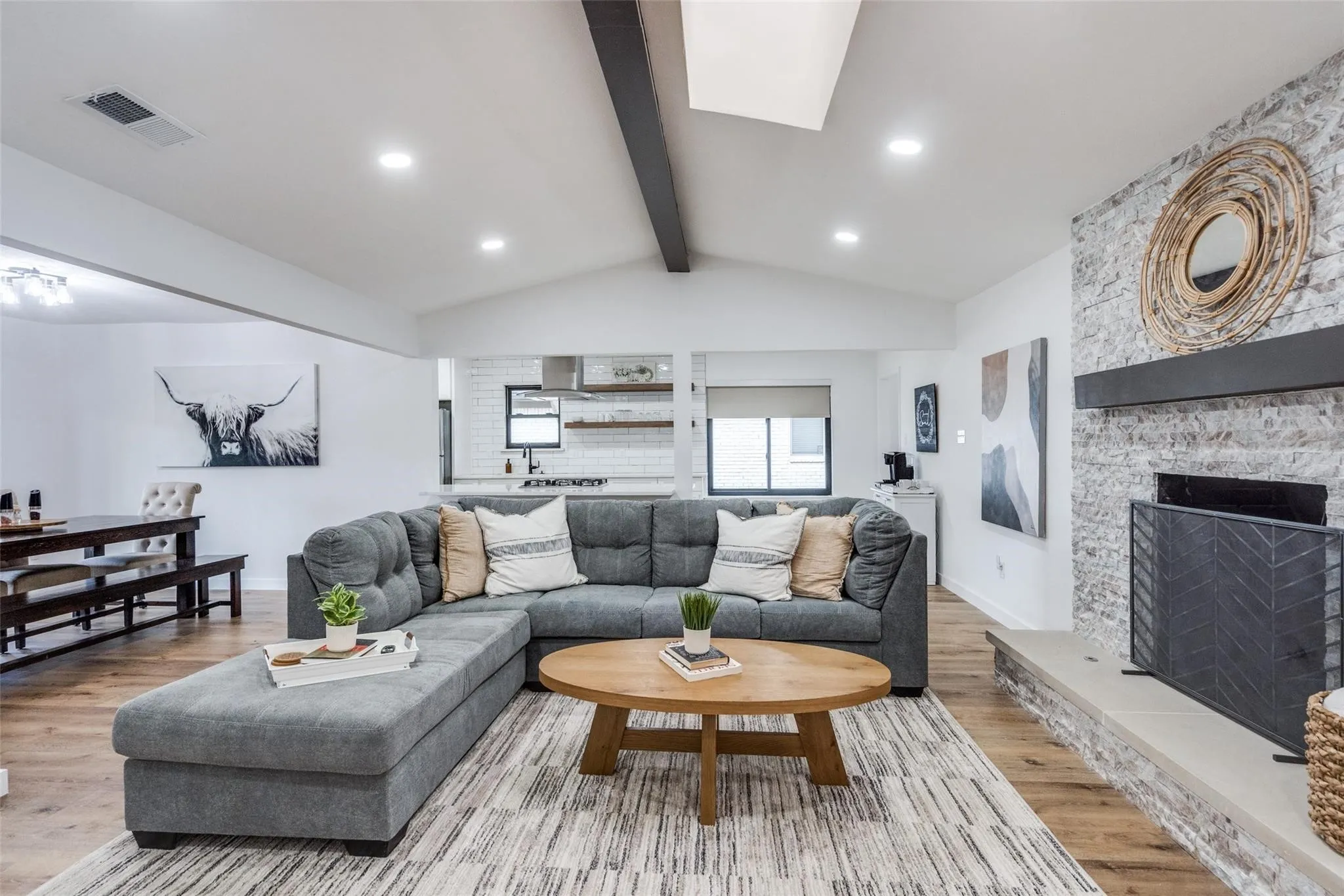 Vaulted ceiling living area featuring a stone-clad fireplace with mantel, recessed lighting, wood-finish flooring, and an open concept layout connecting to the kitchen