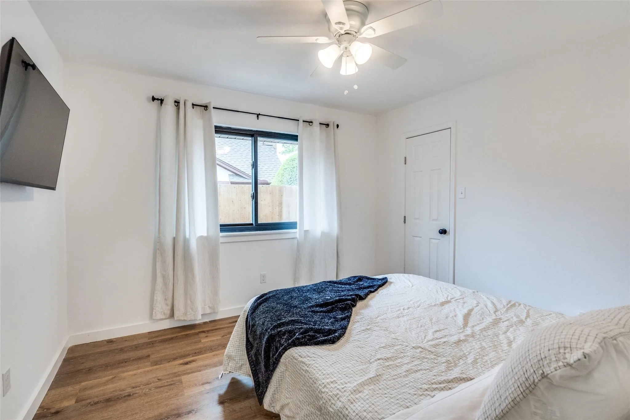 Bedroom featuring wood-finish flooring, a ceiling fan with integrated lighting, a single window with dark framing, and a white paneled door