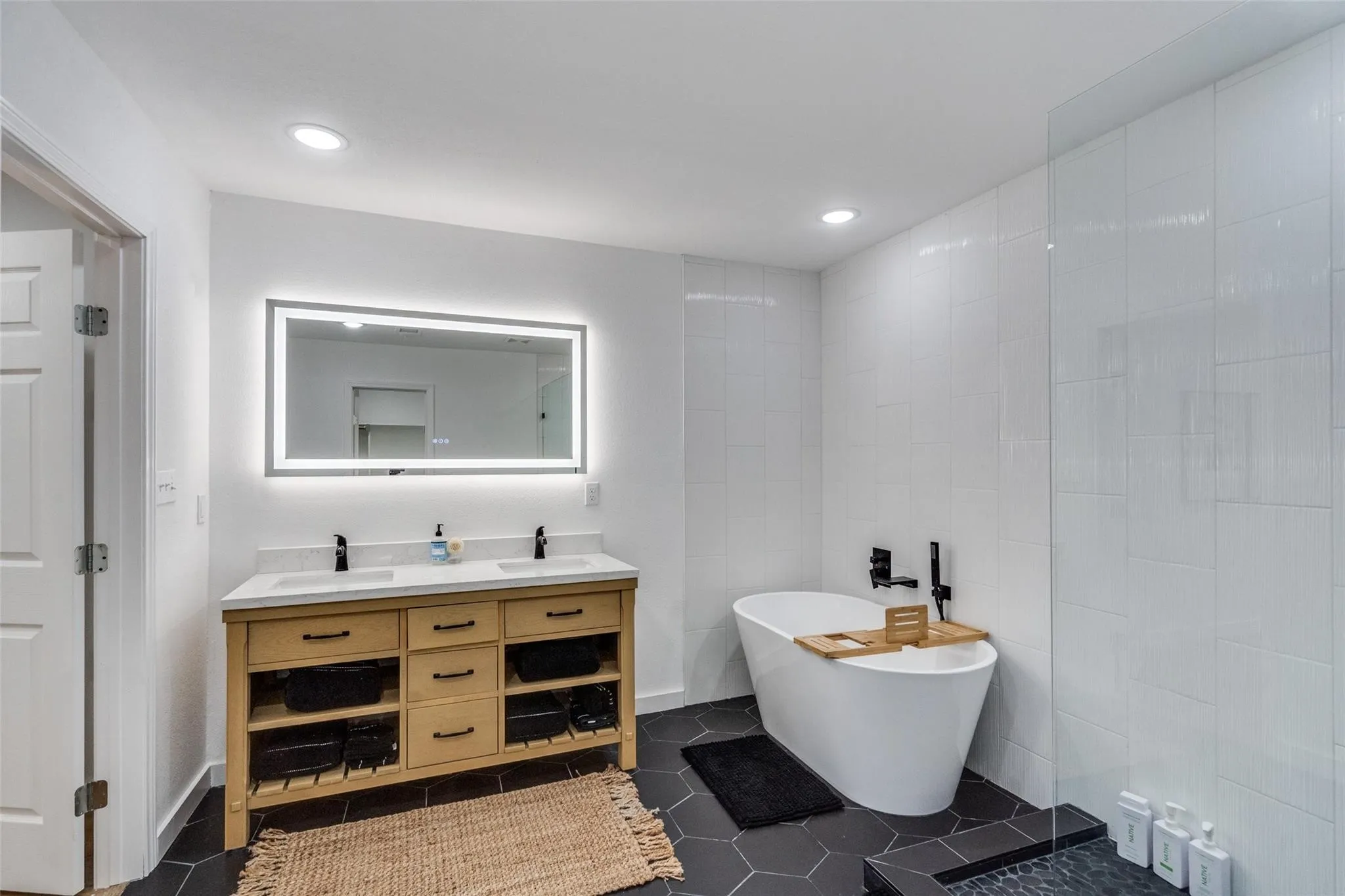 Contemporary bathroom featuring a freestanding soaking tub, a dual vanity with light wood cabinetry, a lighted mirror, and hex tile flooring