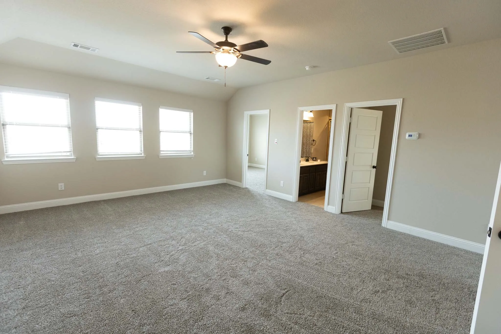 Upstairs Spacious room featuring neutral carpeting, three windows with blinds, a ceiling fan with integrated lighting, and multiple entryways including a visible bathroom vanity with dark wood-finish cabinetry
