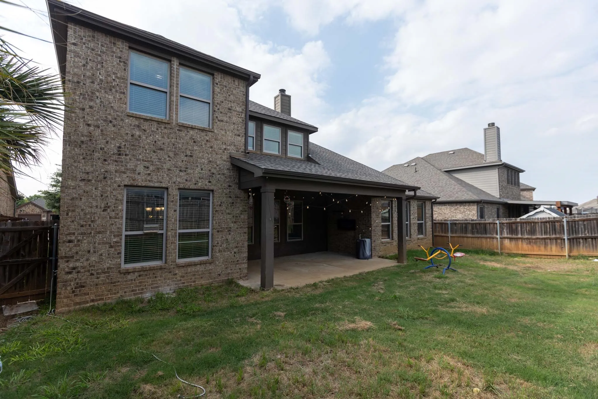 Two-story brick exterior featuring an expansive covered patio, multiple windows with blinds, a fenced yard, and a chimney