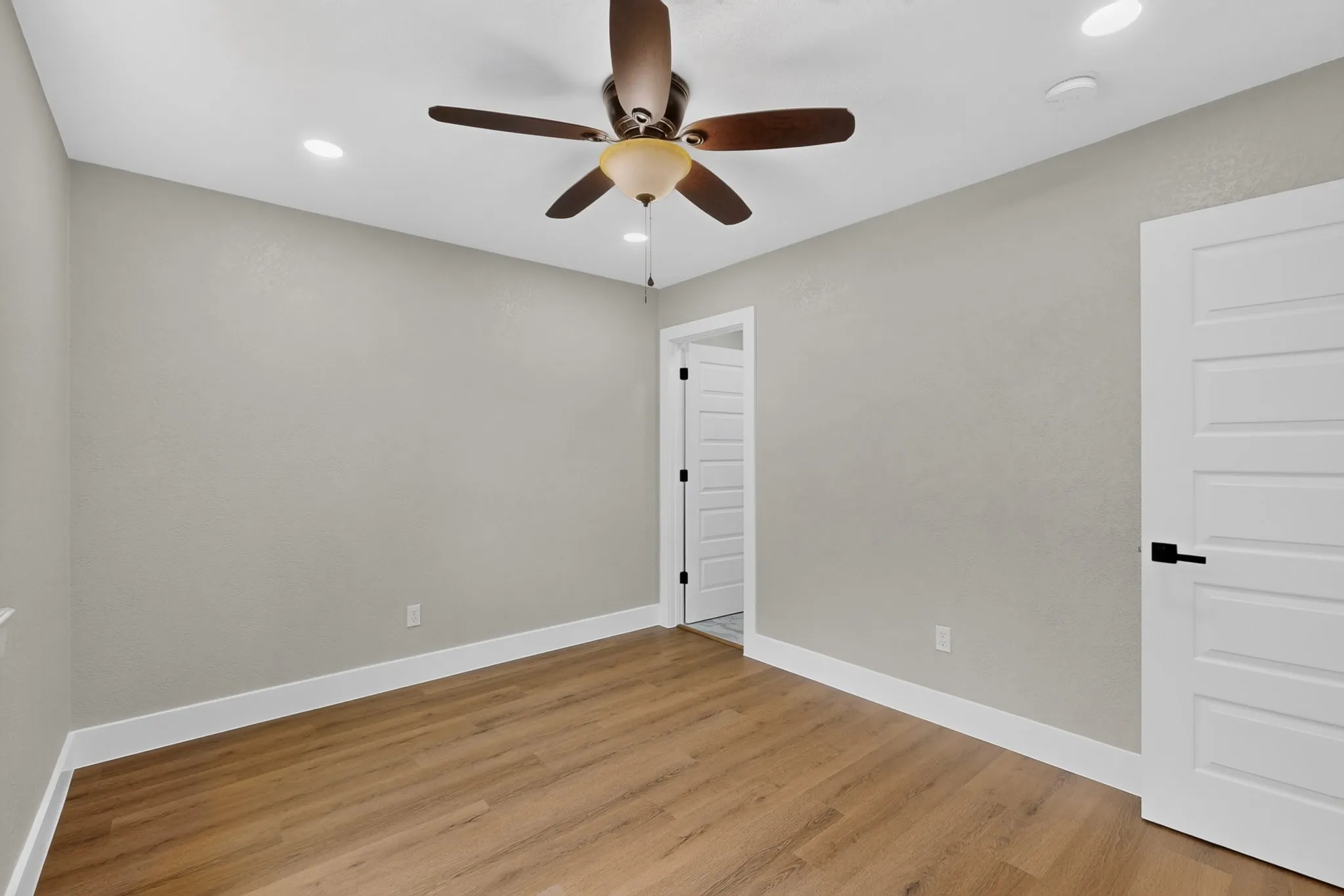 Wood-finish flooring complemented by white baseboards, a ceiling fan with integrated lighting, recessed ceiling lights, and panel doors with matte black hardware