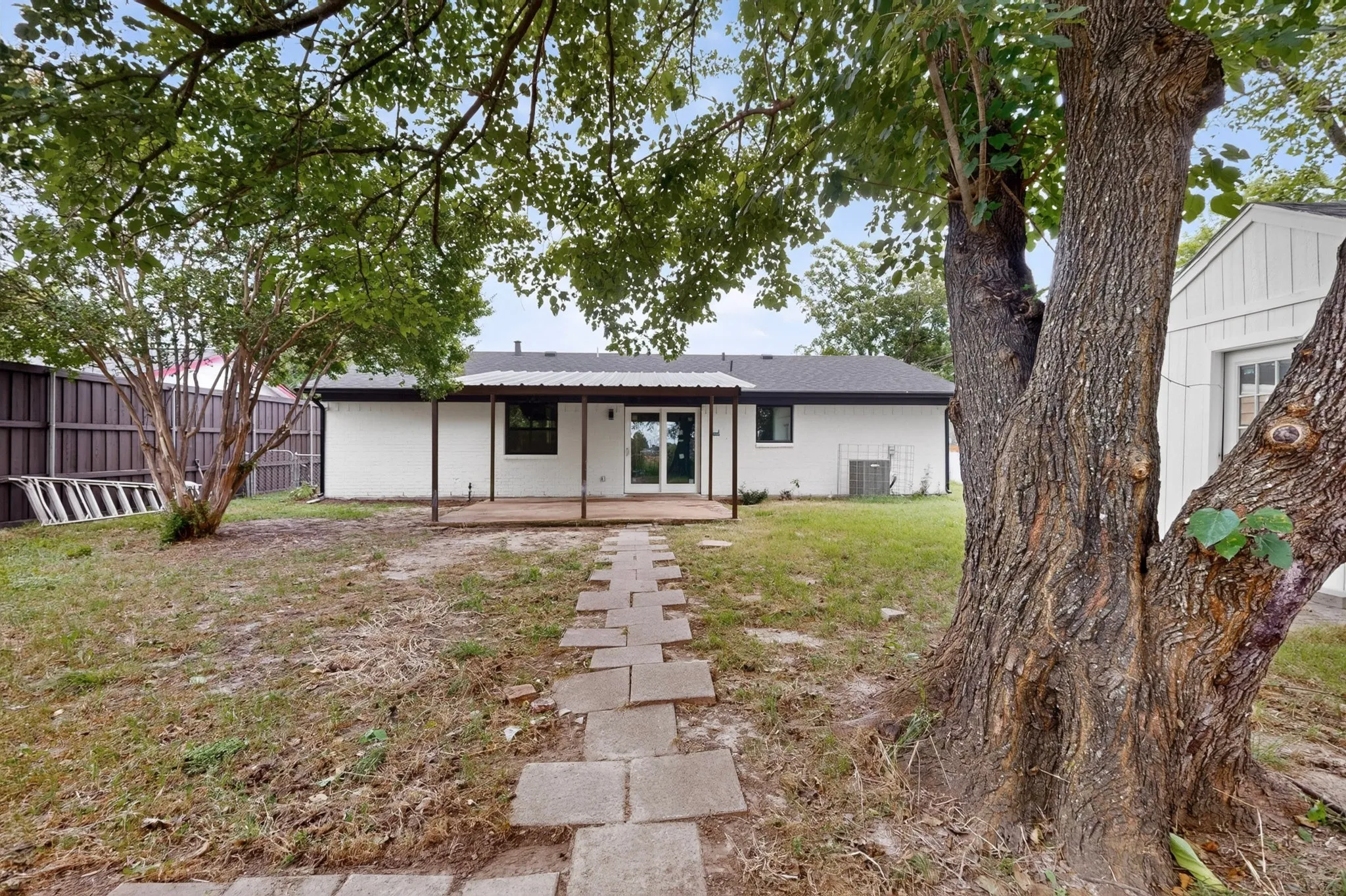 Spacious backyard featuring mature trees, a stone paver pathway, and a covered back patio with support columns