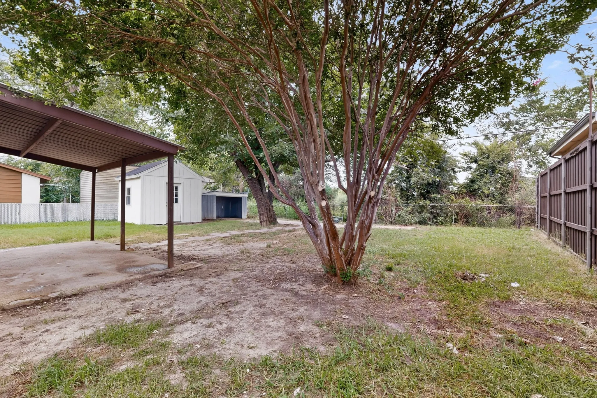 Expansive outdoor space featuring a covered carport, two separate storage sheds, a mature tree, and perimeter fencing