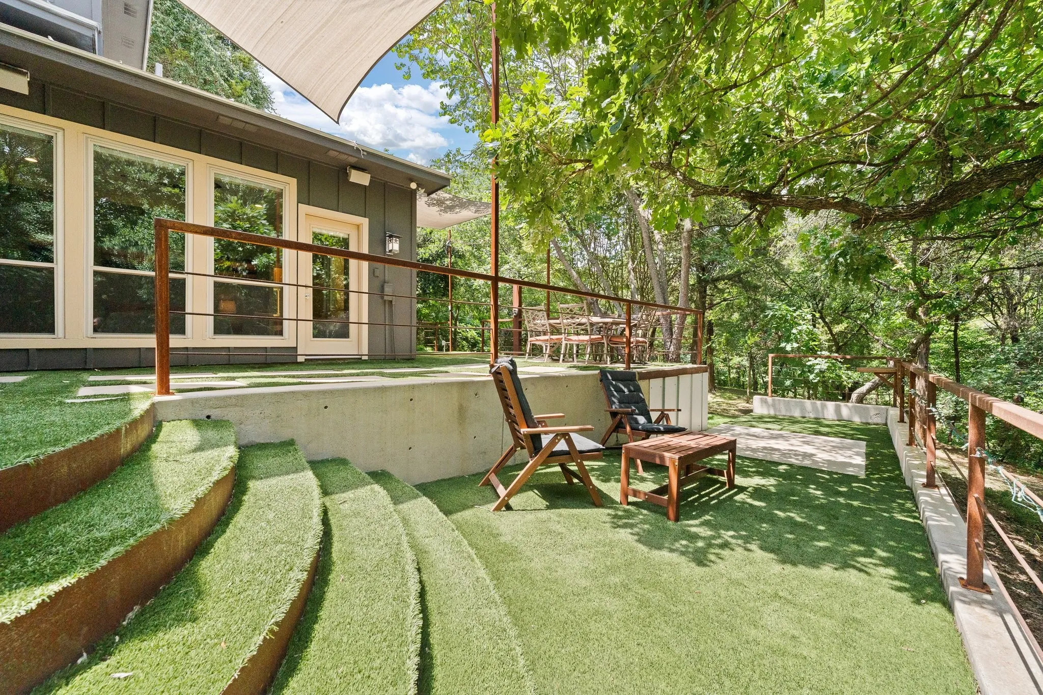 The expansive wraparound patio area offers multi-level outdoor space featuring artificial turf throughout,  metal railings for safety, tiered landscaping, and mature trees providing natural coverage, this sitting area offers a under storage as well and concrete patio area for grilling