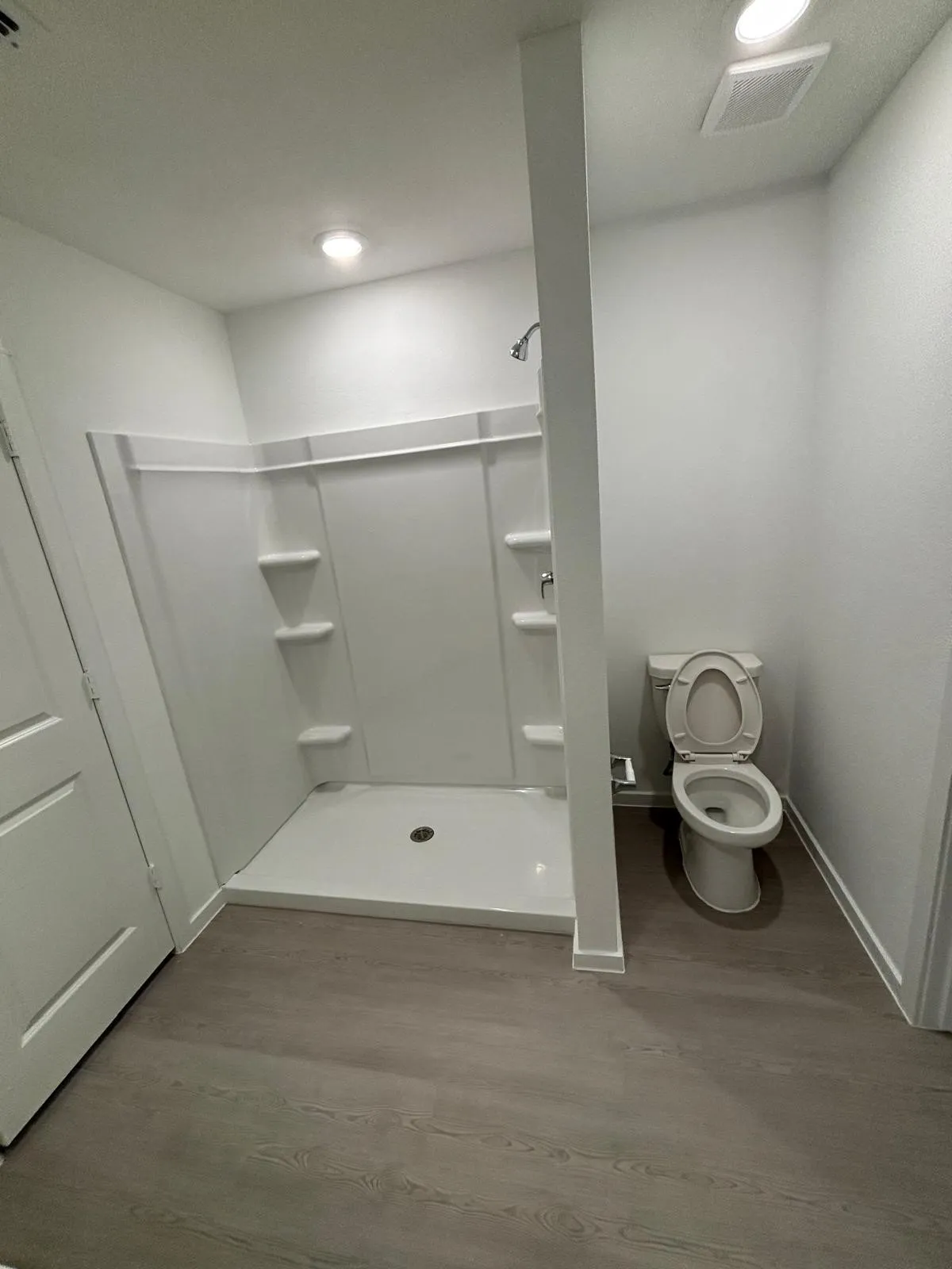 Full bathroom featuring a white stall shower with integrated shelving, a white toilet, and wood-finish flooring