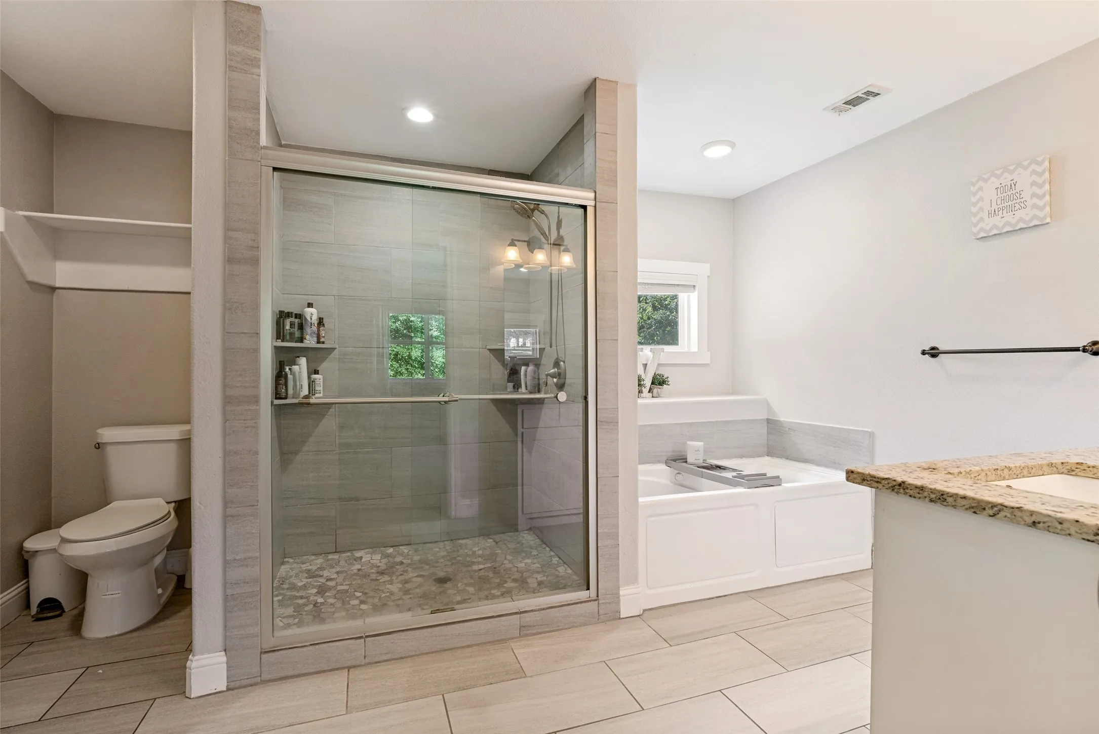 Spacious bathroom featuring a glass-enclosed shower with pebble flooring and built-in shelving, a soaking tub, and a vanity with a granite countertop and integrated sink