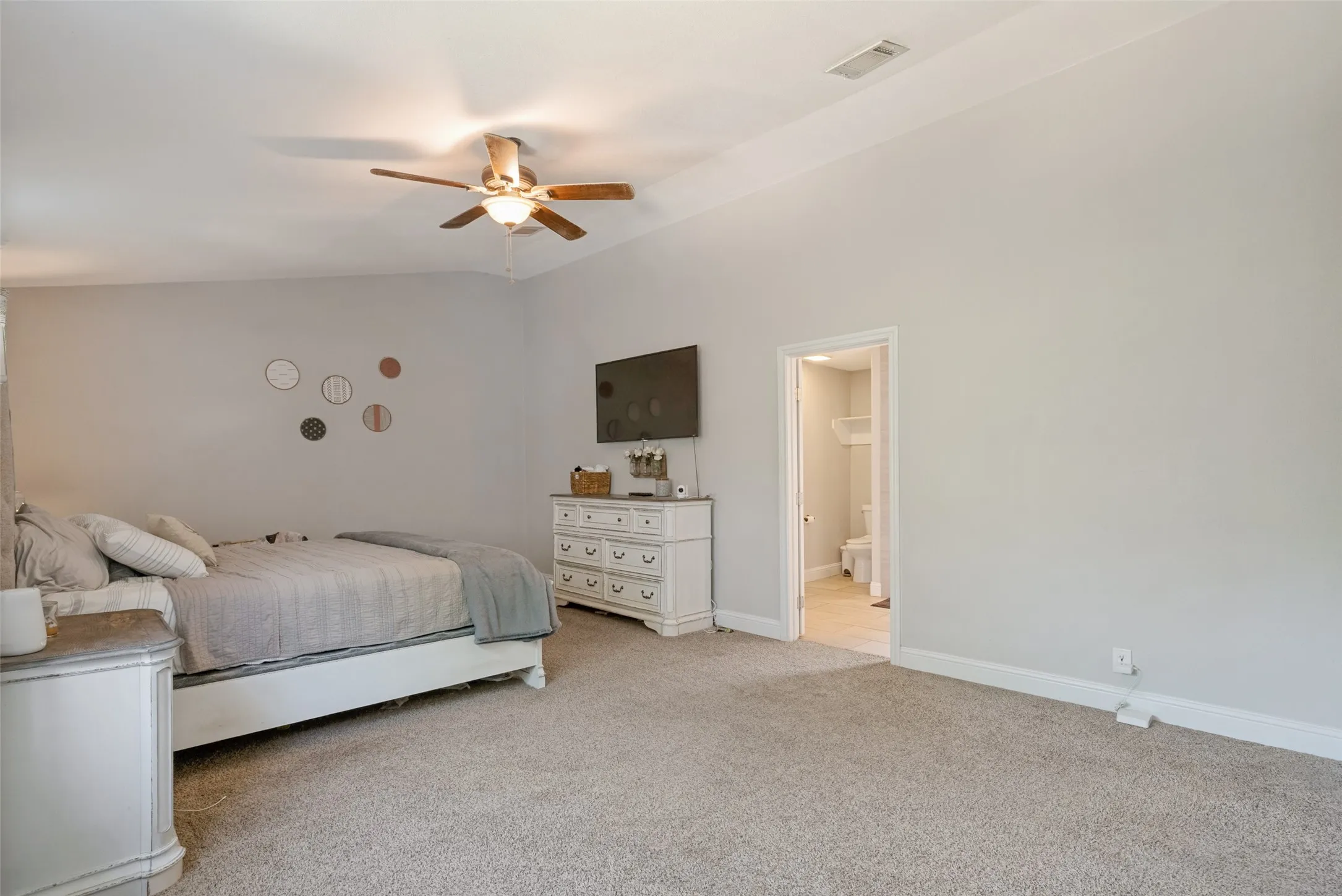 Spacious sleeping quarters featuring light gray wall paint, textured carpeting, a ceiling fan with integrated lighting, and an entryway to an adjoining bathroom with tile flooring