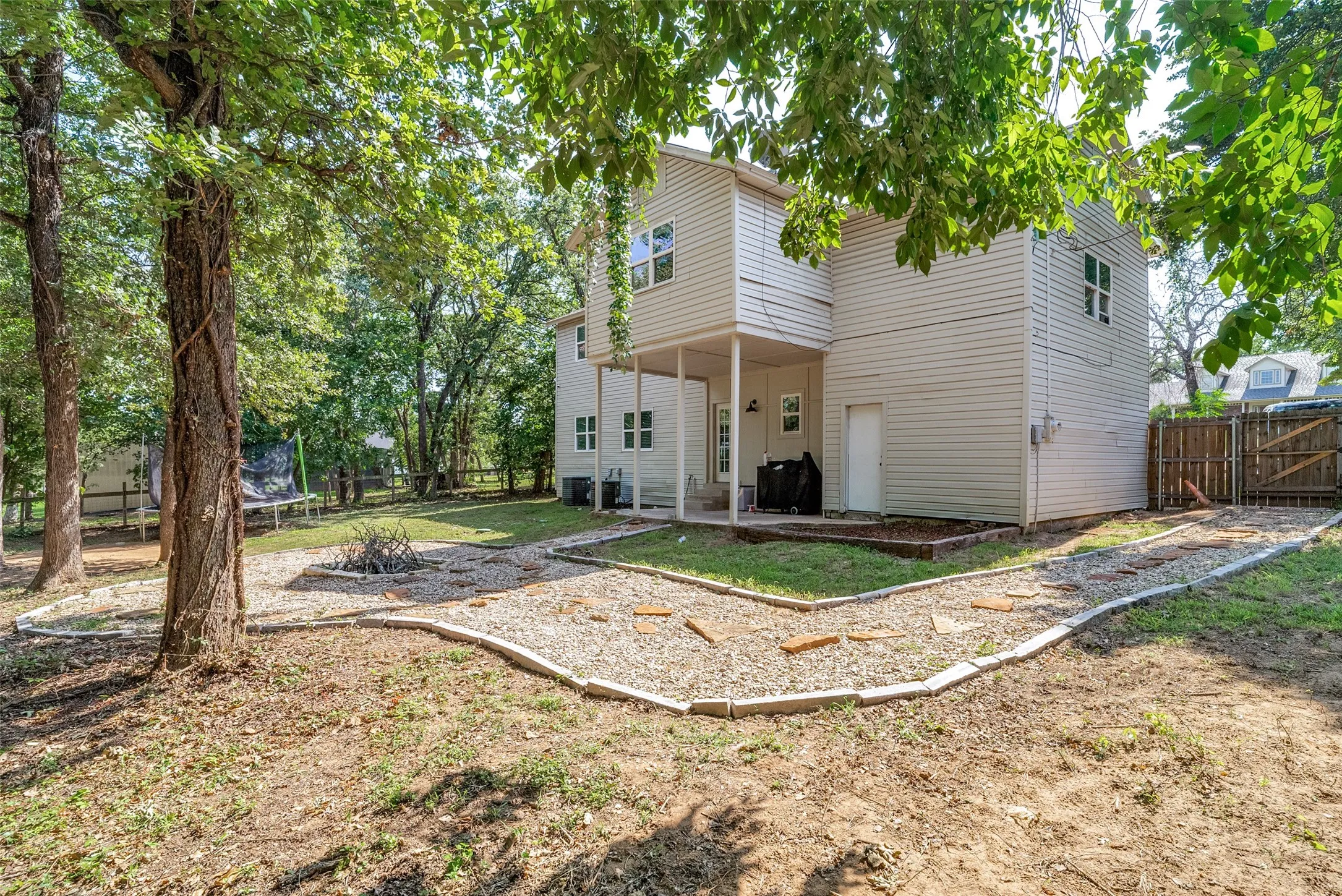 Spacious backyard featuring mature trees, a gravel pathway with stepping stones, and a dedicated fire pit area