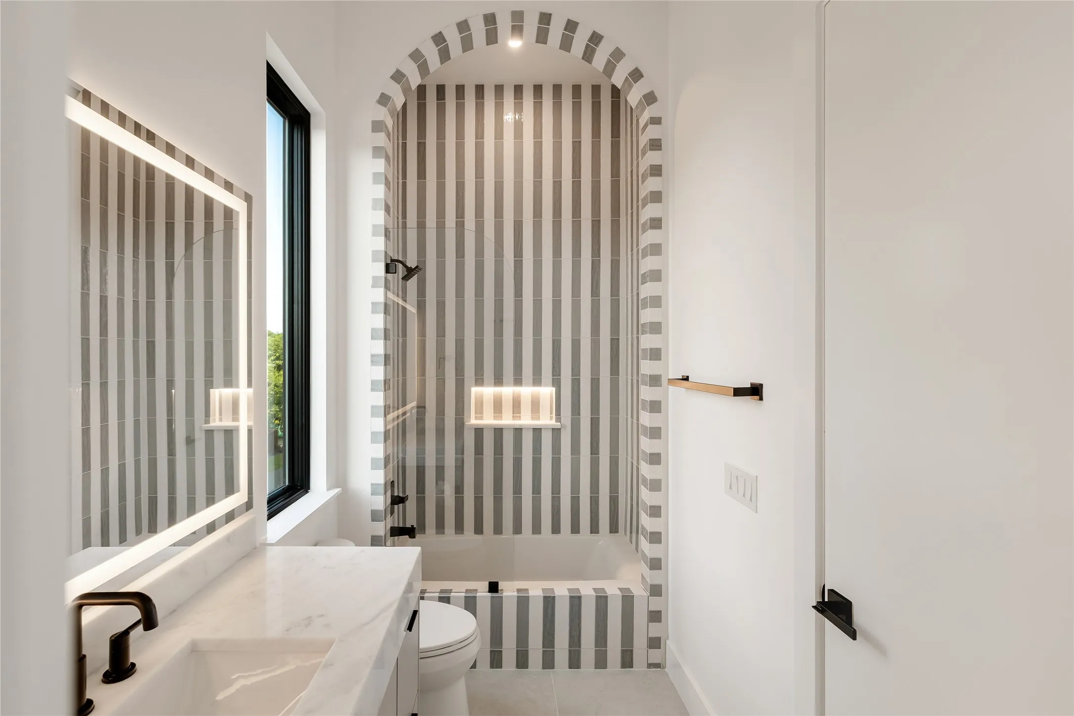 Bathroom featuring a striped tile tub/shower surround with an arched entry, a built-in illuminated niche, a contemporary vanity with an integrated sink, a rectangular backlit mirror, and matte black fixtures