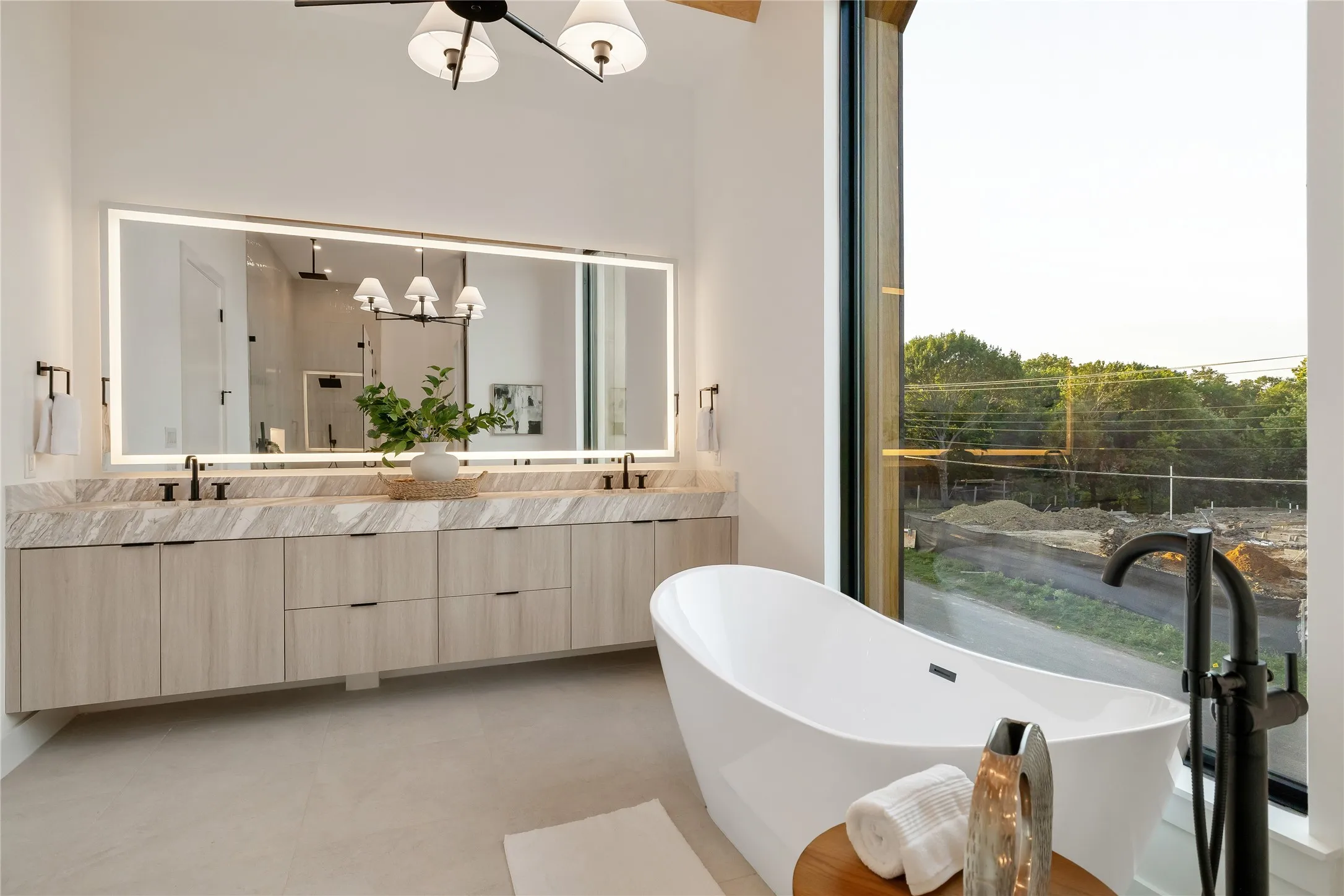 Modern bathroom featuring a freestanding soaking tub with floor-mounted fixture, a large vanity with dual sinks and light wood-finish cabinetry, a full-wall illuminated mirror, and a floor-to-ceiling window