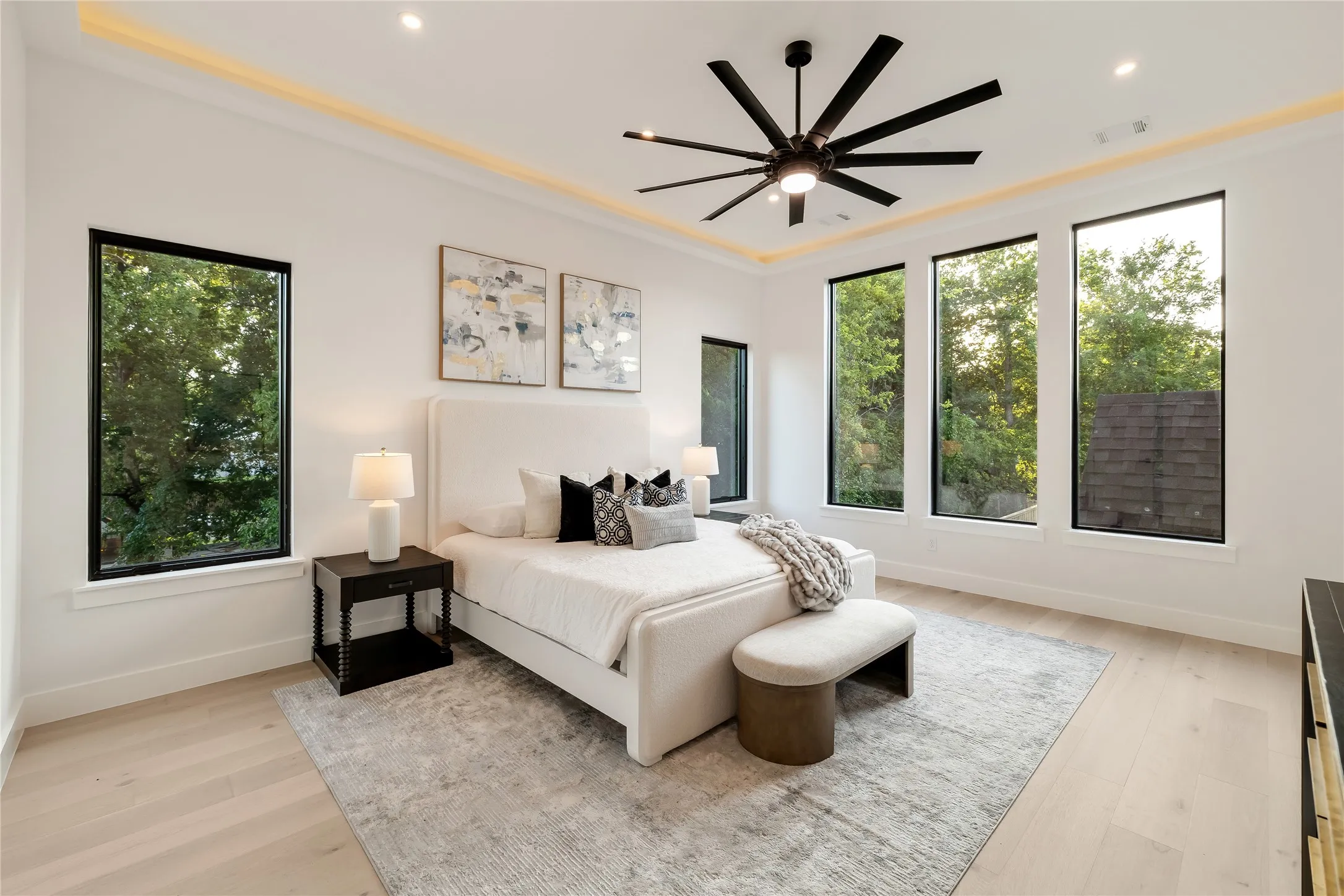 Bright interior space with light wood-finish flooring, featuring four tall windows with black frames, a contemporary ceiling fan, and recessed lighting with perimeter accent lighting