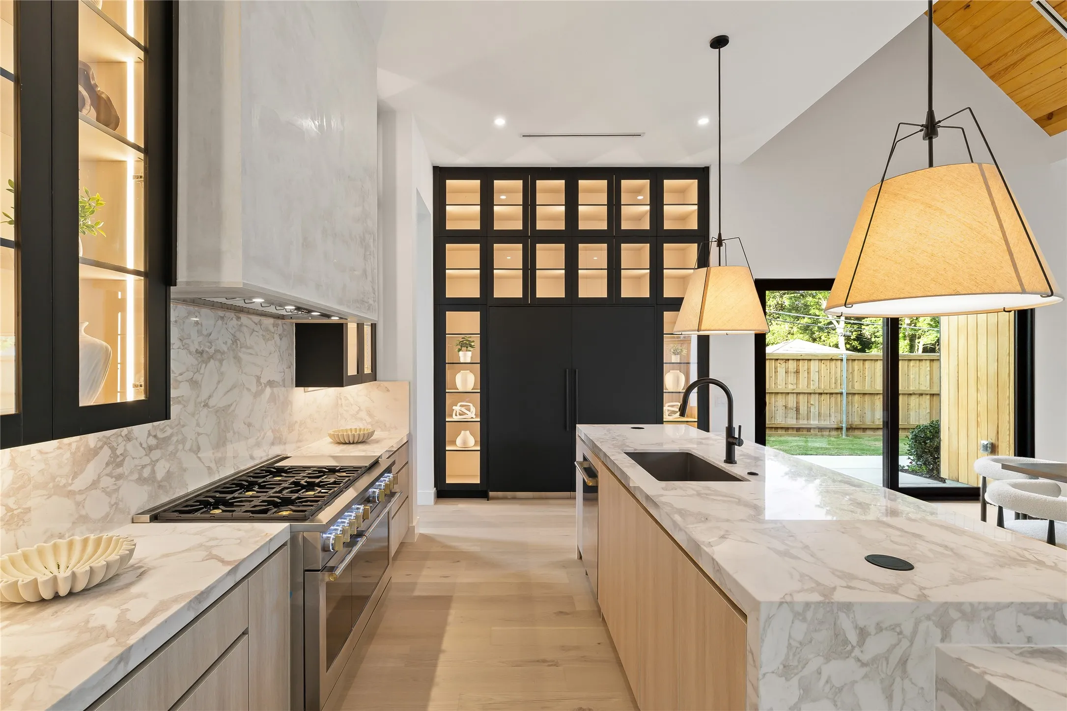 Gourmet kitchen featuring a substantial island with a built-in sink and matte black faucet, integrated stainless steel appliances, light wood-finish cabinetry, extensive marbled countertops and backsplash, and a contemporary range hood