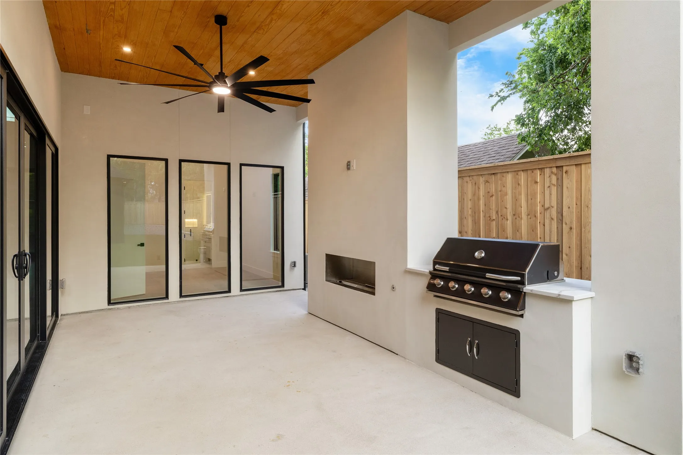 Covered patio featuring a wood-paneled ceiling with a modern ceiling fan, an outdoor kitchen with a built-in grill, stone countertop, and integrated storage, and large glass panel doors