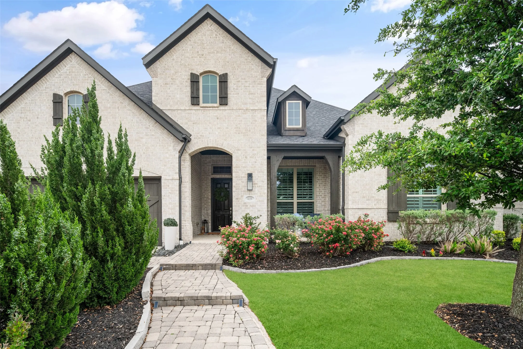 A beautiful brick paver walkway leads to the covered front porch, surrounded by professionally designed landscaping with lush greenery, mature trees, vibrant flowering shrubs, and impeccably manicured lawns, creating exceptional curb appeal and a warm, welcoming entrance.