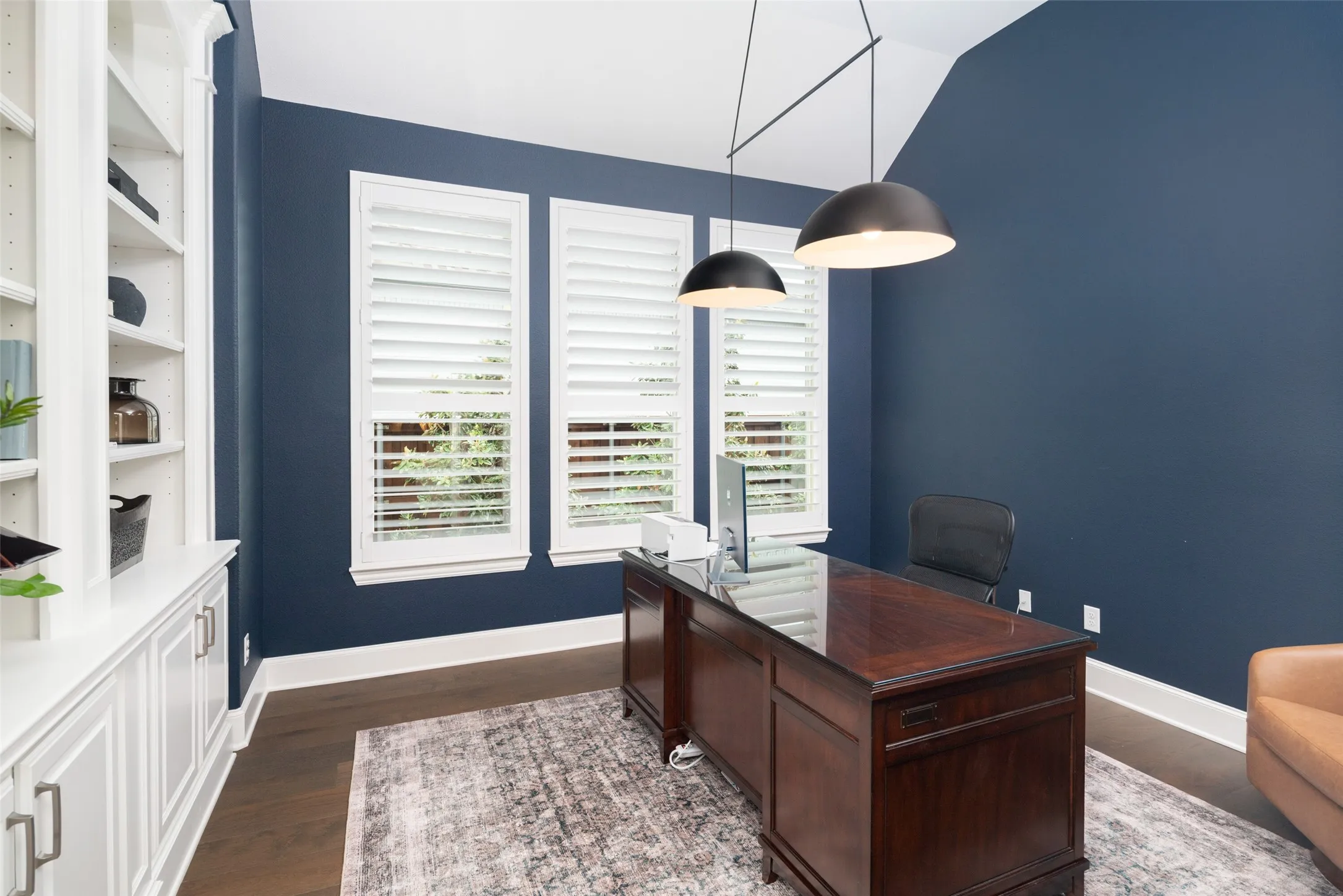 A private home office blends style and functionality with three large windows adorned with plantation shutters, filling the space with abundant natural light. Rich wide plank wood floors, a designer upgraded light fixture, and custom built-in book shelving create a refined workspace that's ideal for working from home or a quiet study.