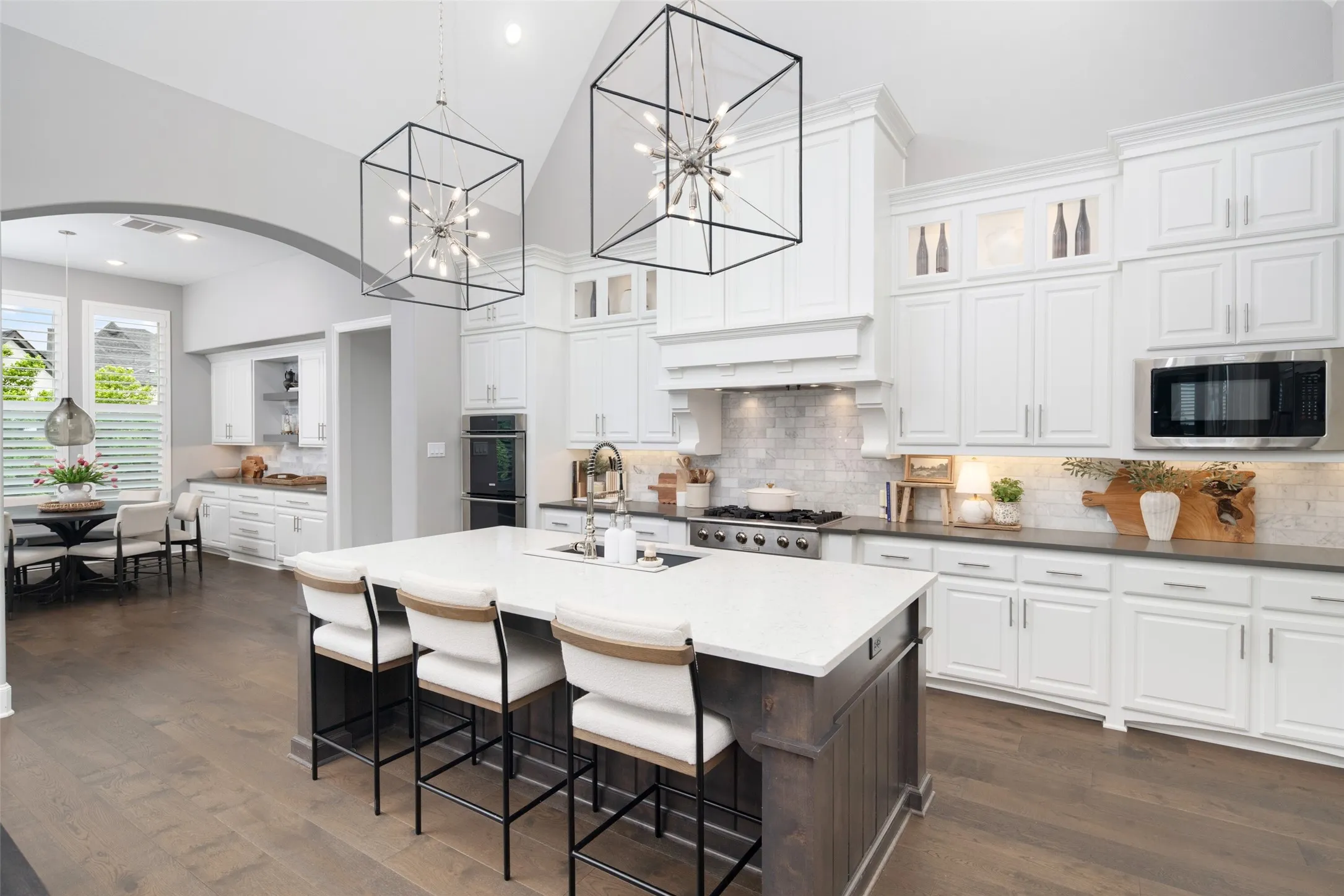 The heart of the home is a breathtaking chef's kitchen showcasing soaring ceilings, dramatic designer statement light fixtures, striking two-tone cabinetry, an oversized quartz island with seating, expansive quartz countertops, a designer tile backsplash, and upper glass display cabinets with accent lighting. Thoughtfully designed for both everyday living and effortless entertaining, this stunning space flows seamlessly into the breakfast area and living room.