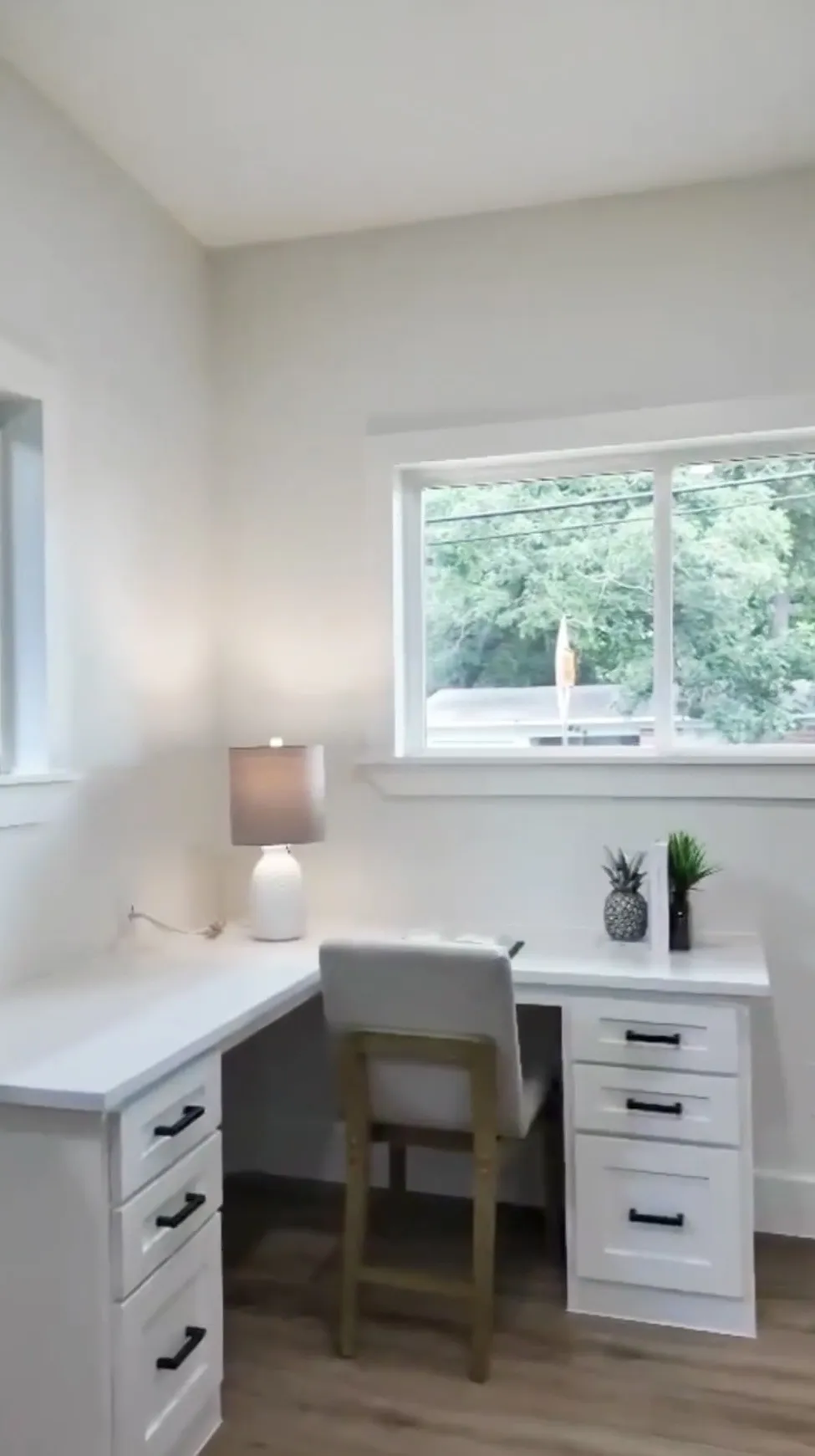 Room featuring built-in L-shaped desk with white cabinetry and dark hardware, two windows with white trim, and wood-finish flooring