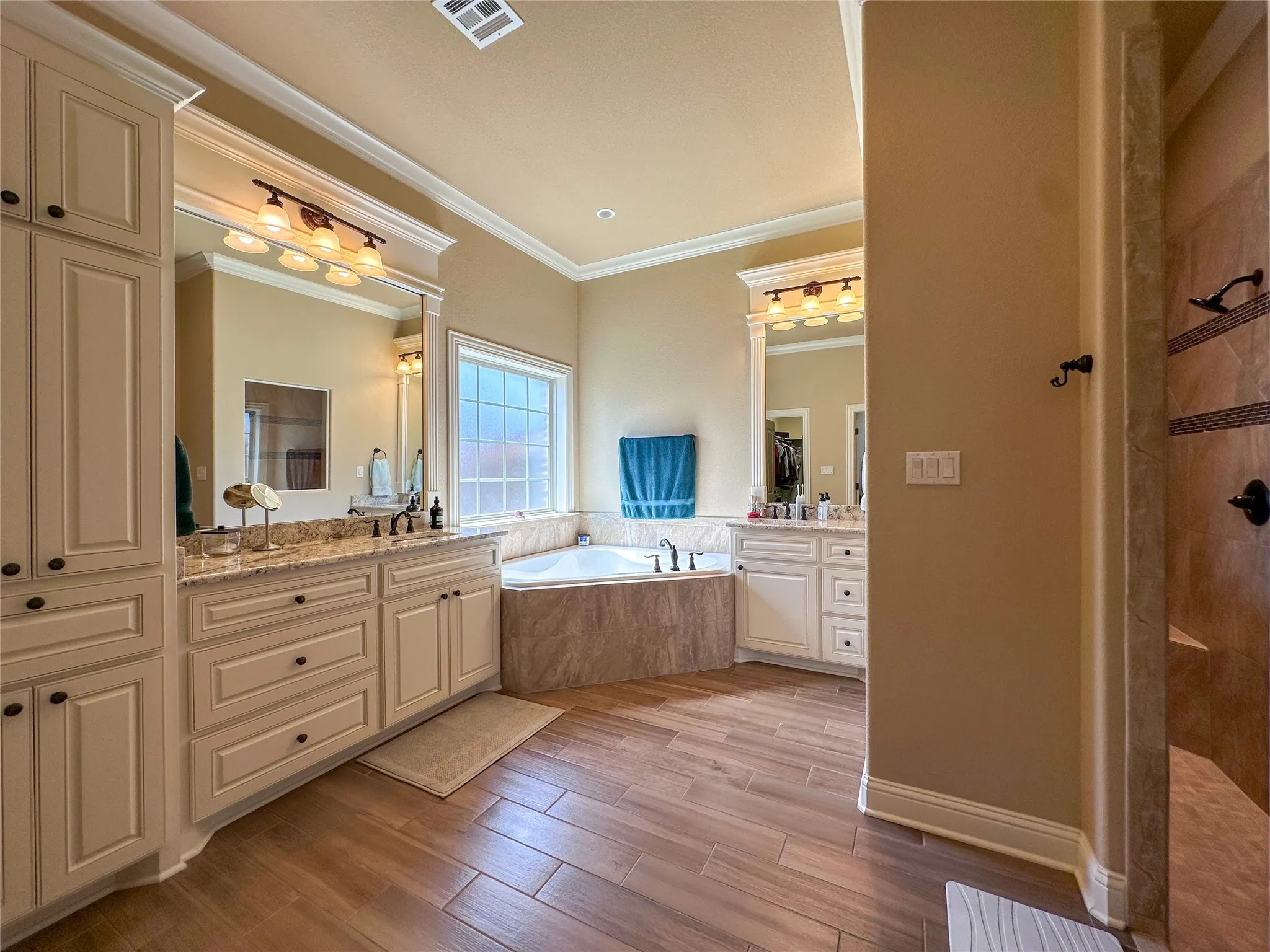 Bathroom featuring wood-finish flooring, a corner soaking tub with tiled surround, and dual vanities with granite countertops