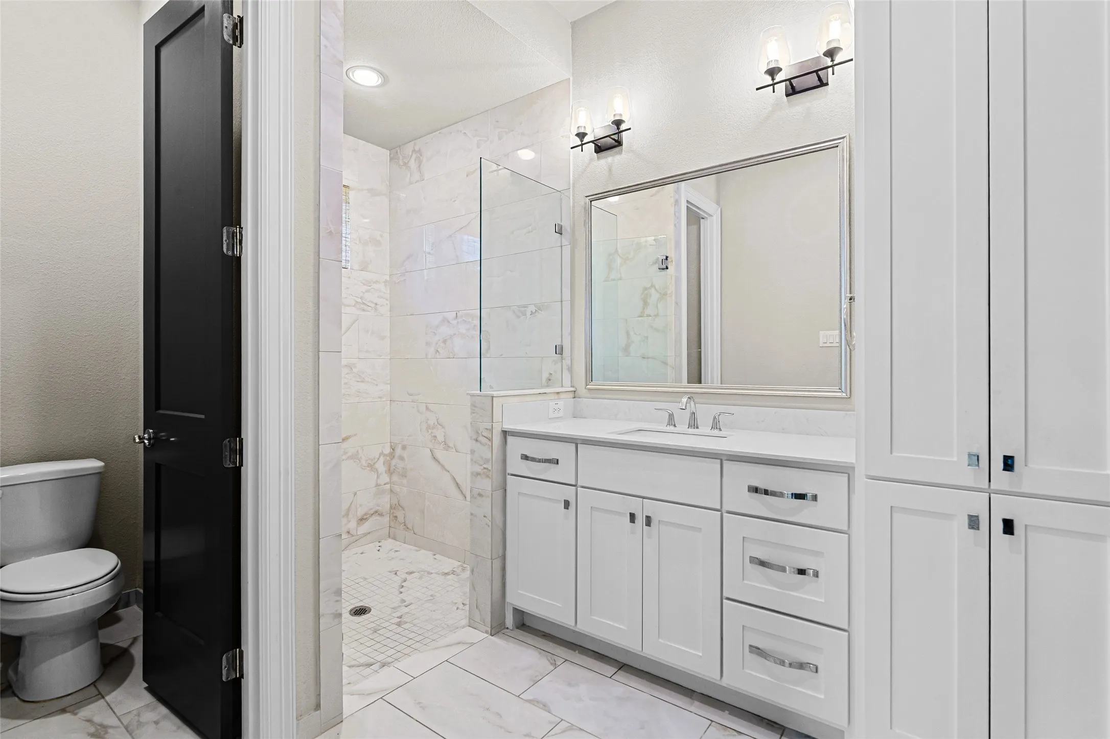Bathroom featuring a white vanity with integrated sink, a frameless glass shower enclosure, white tile flooring, recessed lighting, and wall-mounted sconces. Primary bathroom on the 1st floor.