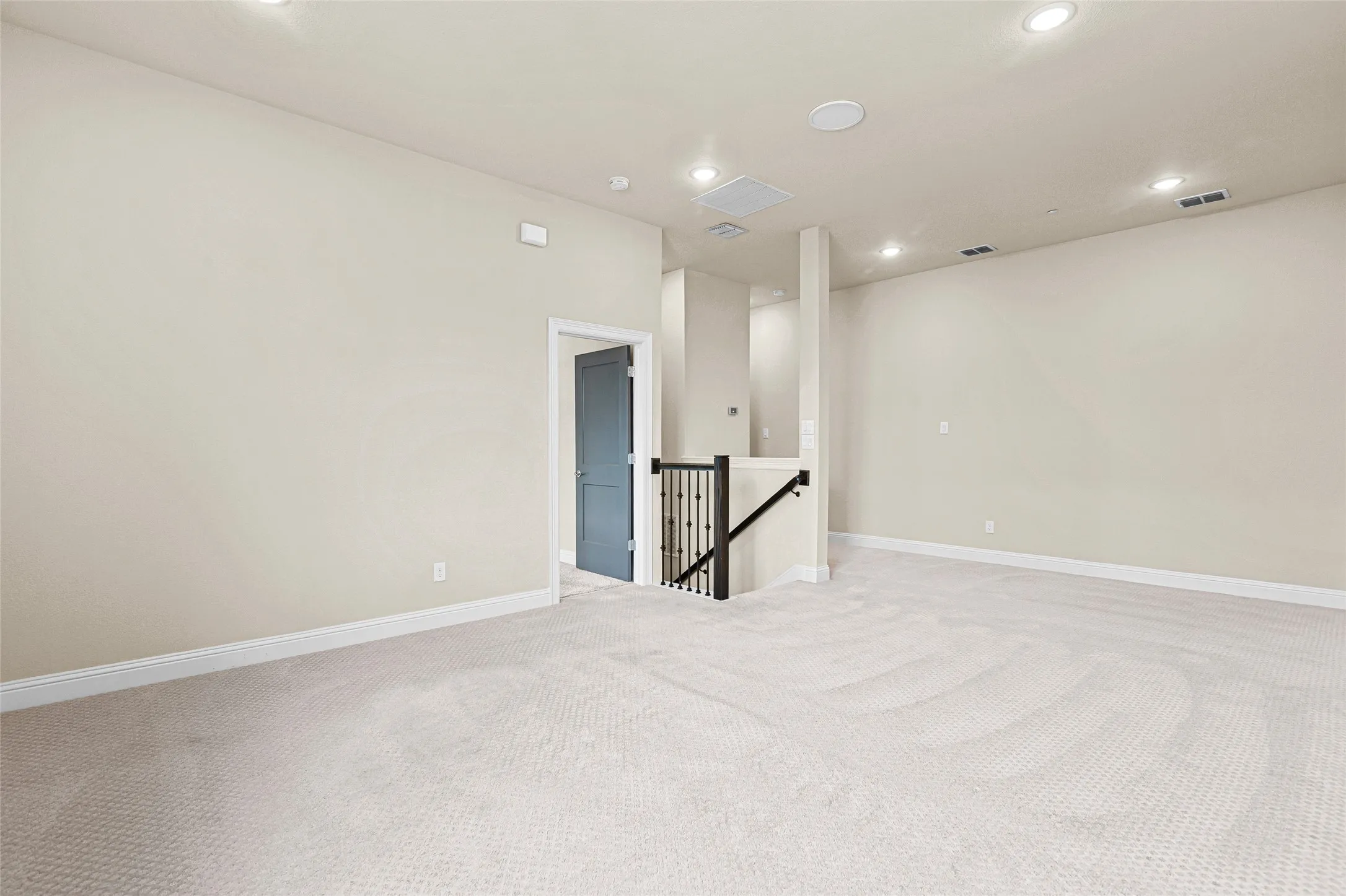 Expansive carpeted room featuring recessed lighting, a neutral color palette, a dark wood and iron railing, and a contrasting interior door