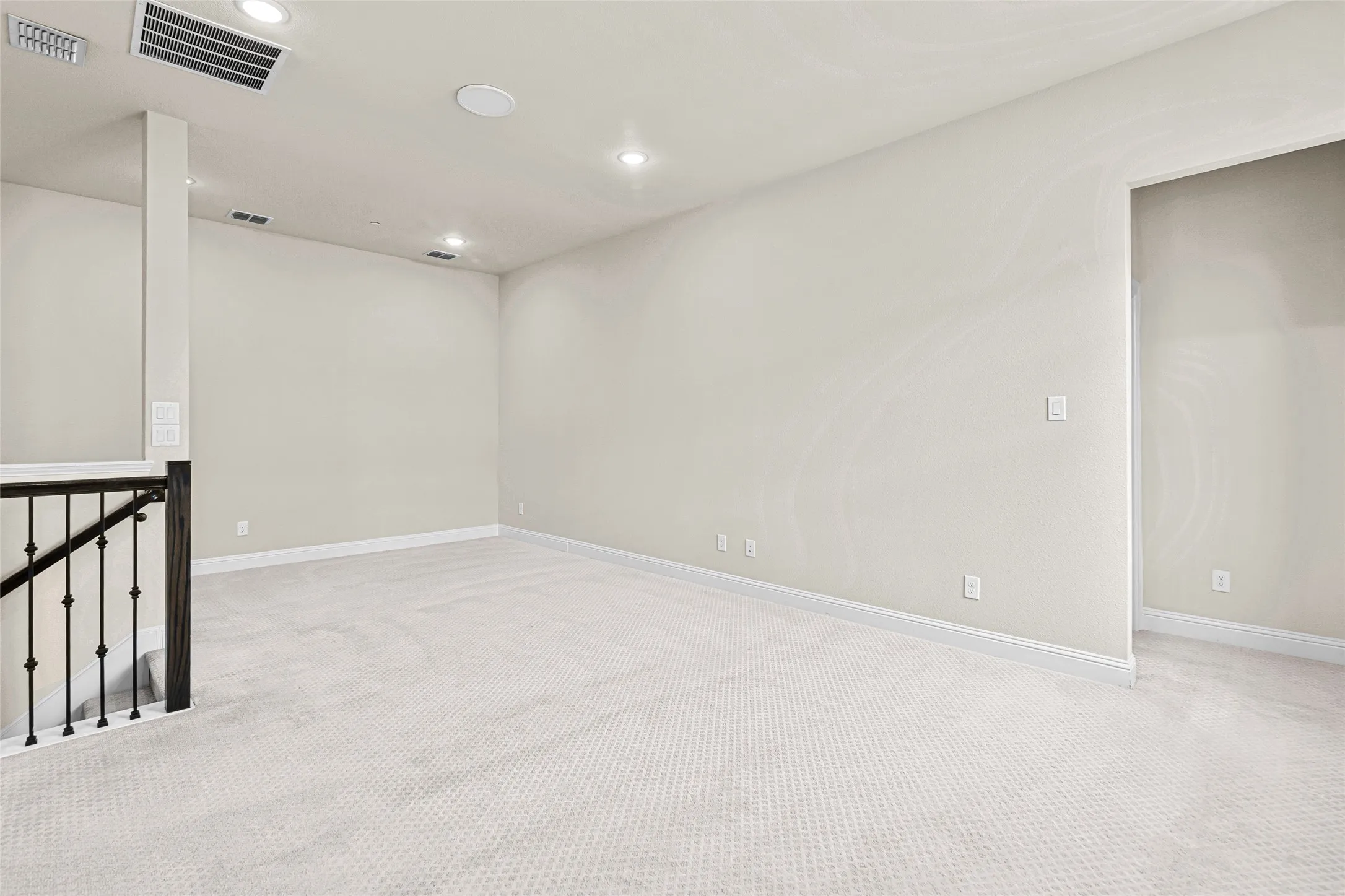 Spacious room featuring light-toned carpeting, recessed lighting, a built-in ceiling speaker, crisp white baseboards, and a dark wood and wrought iron staircase railing