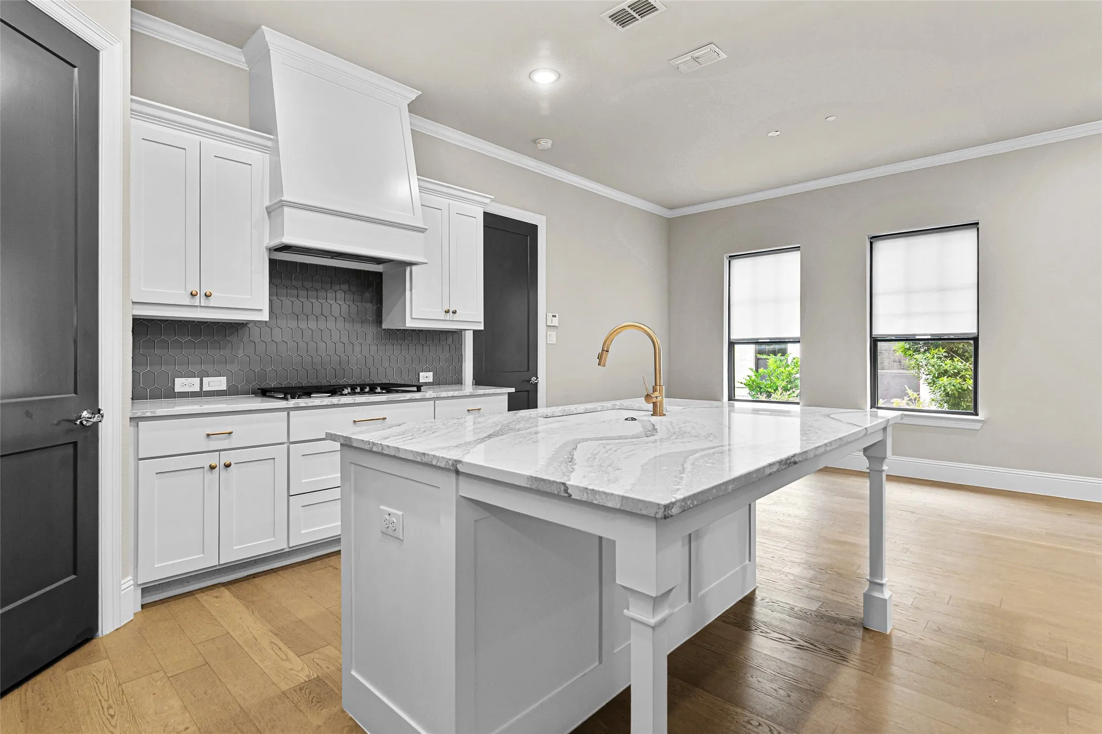 Kitchen featuring a central island with a stone countertop, white shaker cabinetry, a gray hexagonal tile backsplash, a gold-tone faucet, and light wood-finish flooring