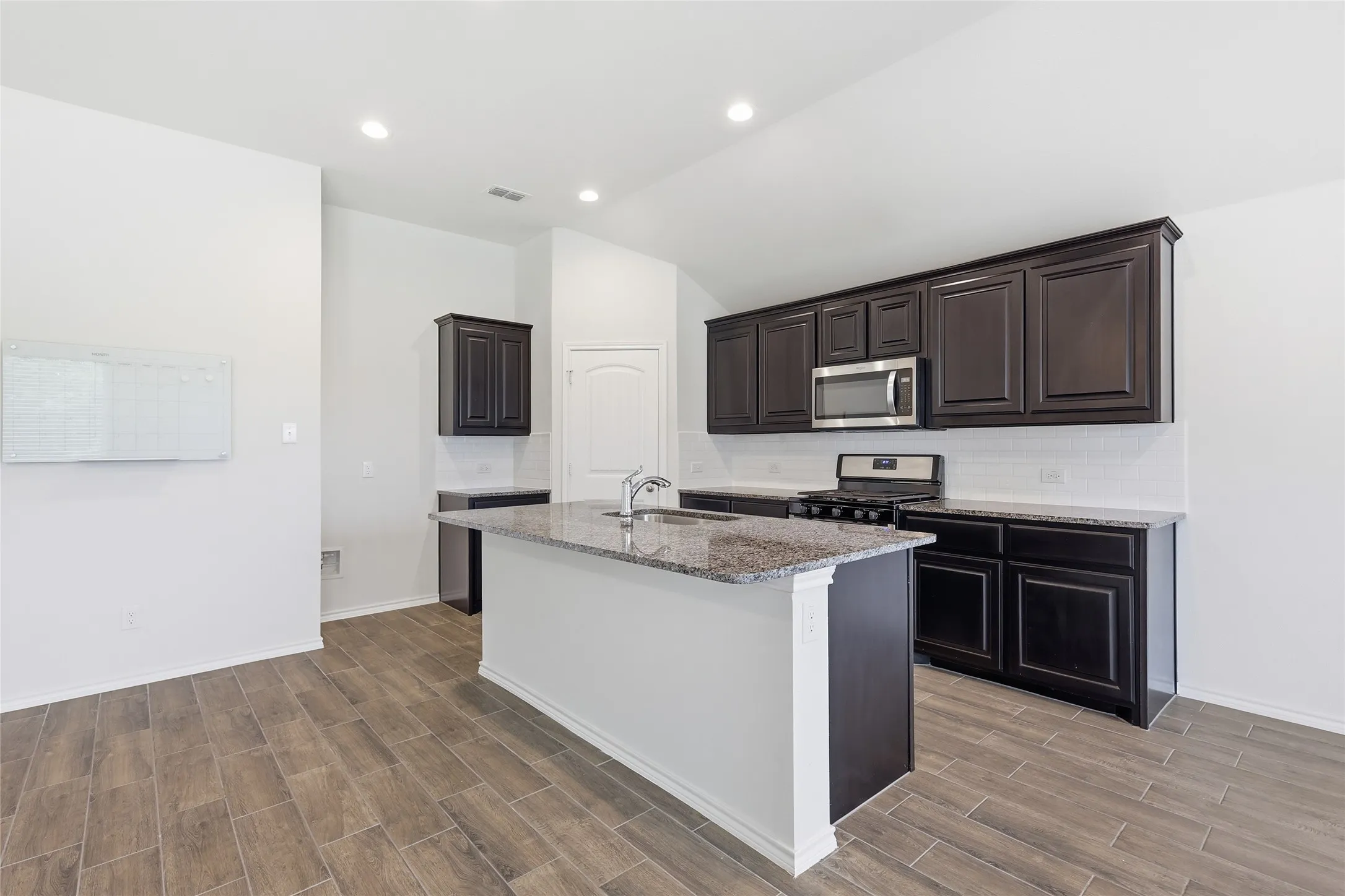 Kitchen featuring an island with a stainless steel sink, granite countertops, dark wood cabinetry, a stainless steel microwave, and wood-finish flooring