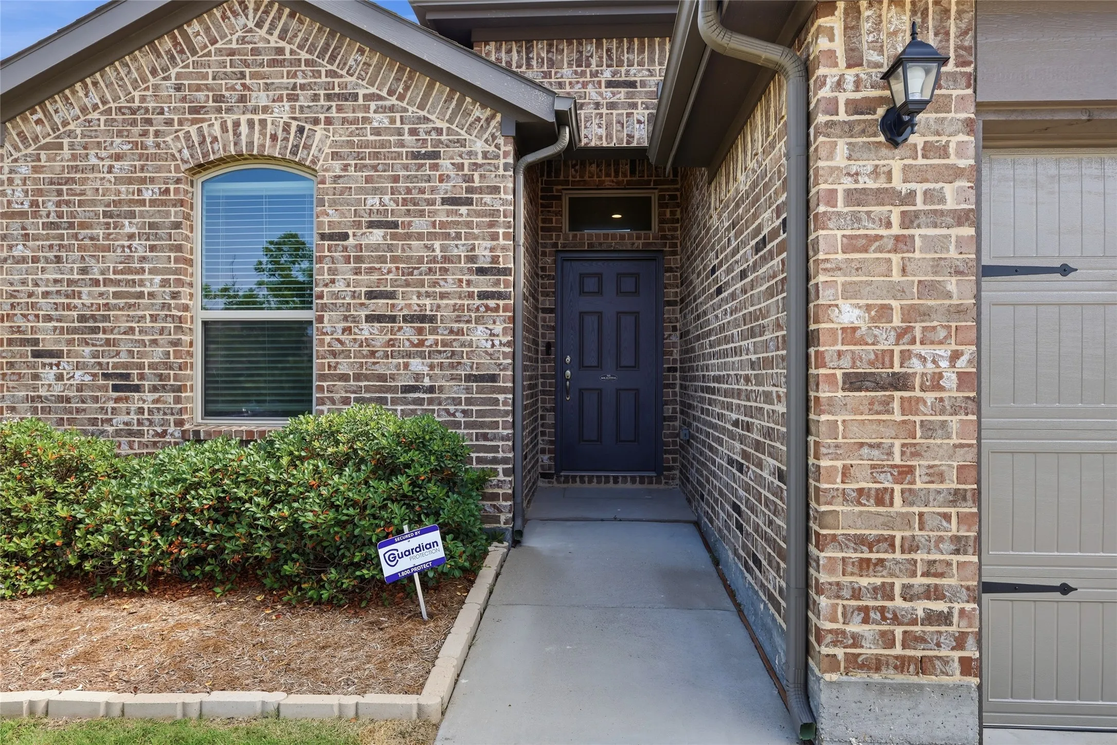 Brick exterior featuring a gray paneled entry door with sidelight, an arched window with blinds, a concrete walkway, and a garage door with decorative hardware
