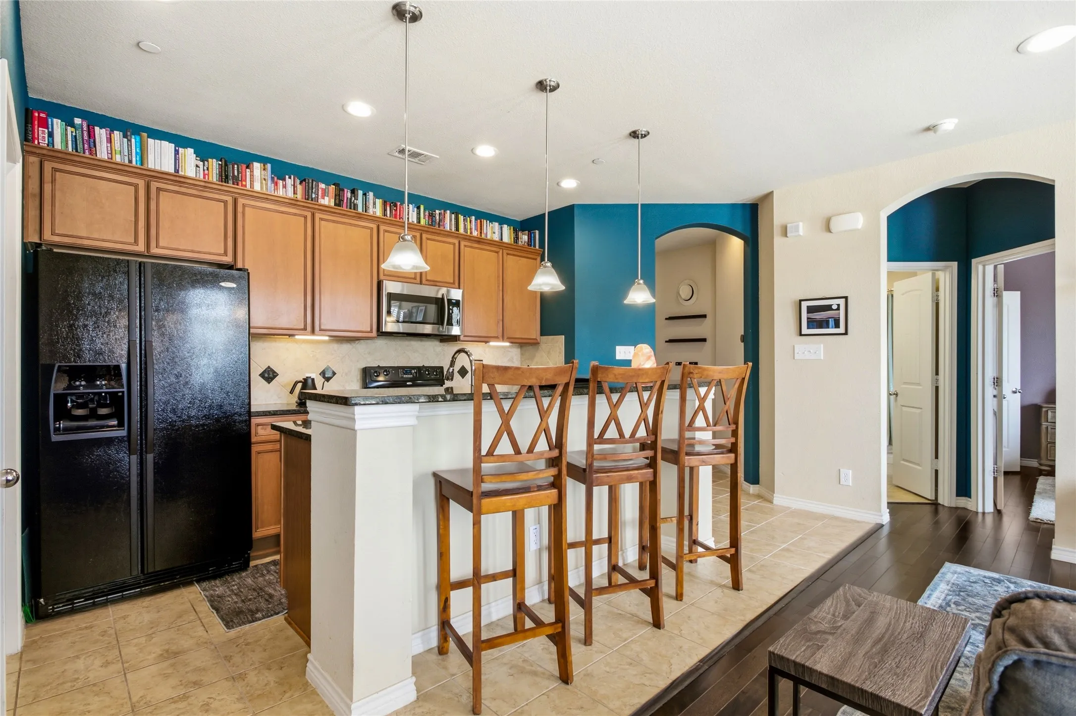 Modern kitchen with wood cabinetry and black appliances, featuring a breakfast bar with pendant lighting, tile flooring, and contrasting dark wood-finish flooring