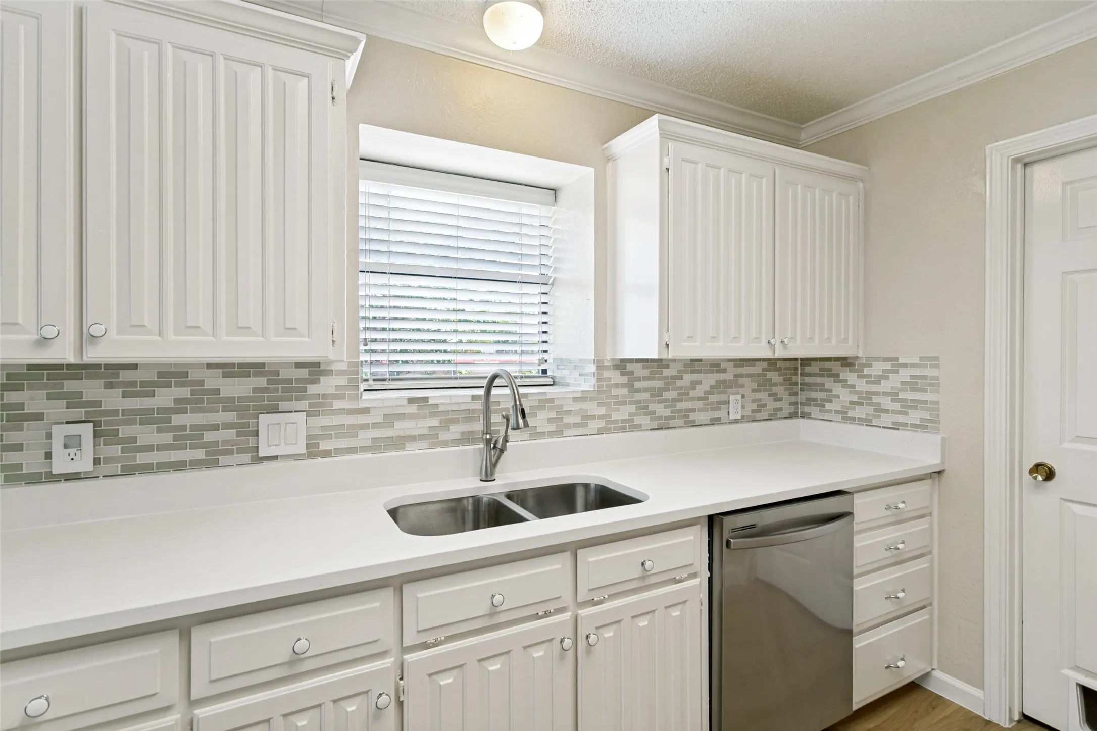 Kitchen featuring white shaker-style cabinetry, a dual-basin stainless steel sink, a stainless steel dishwasher, light-toned countertops, and a mosaic tile backsplash