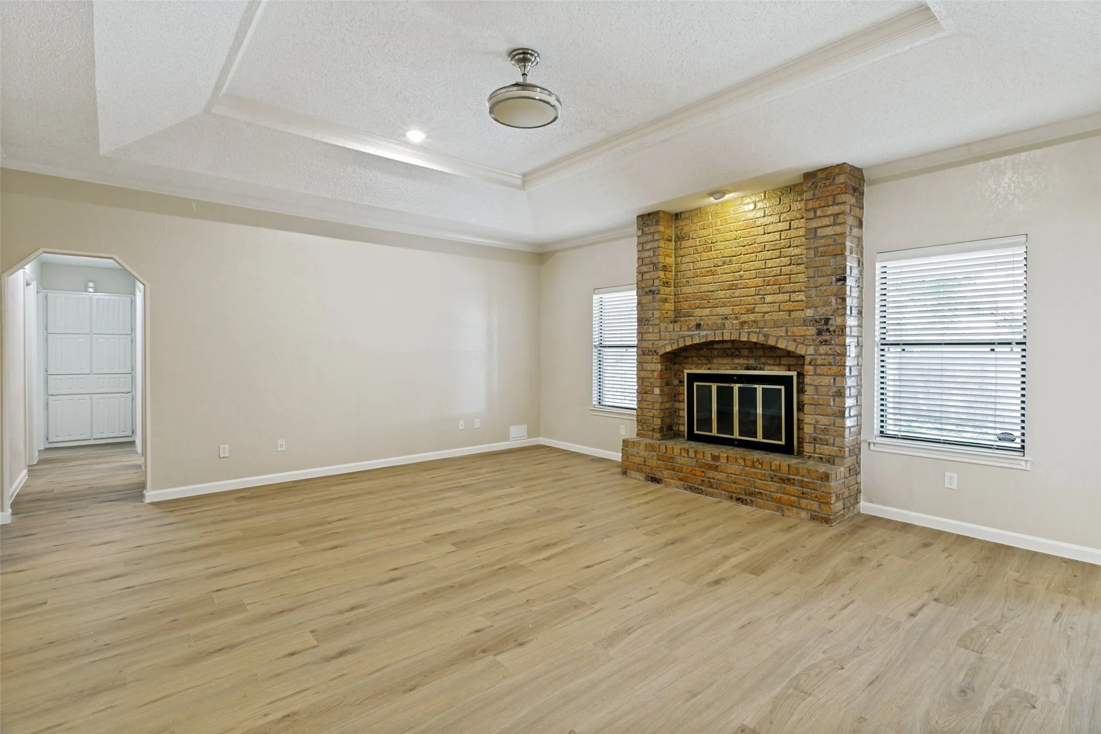 Spacious living area featuring a brick fireplace with a black firebox, wood-finish flooring, and a tray ceiling with a central lighting fixture