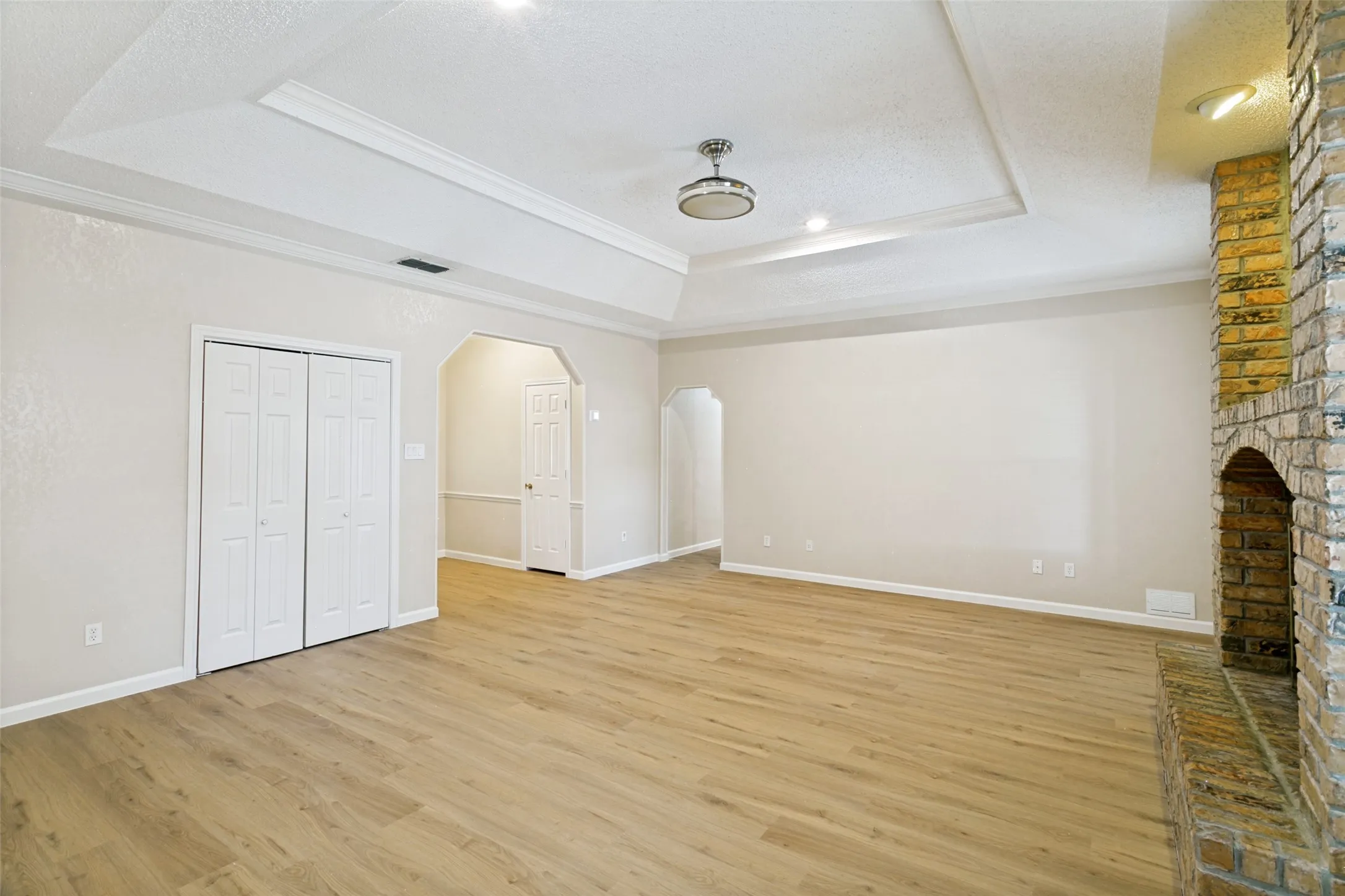 Spacious room featuring wood-finish flooring, a tray ceiling with recessed lighting and a central fixture, a brick fireplace, and arched doorways