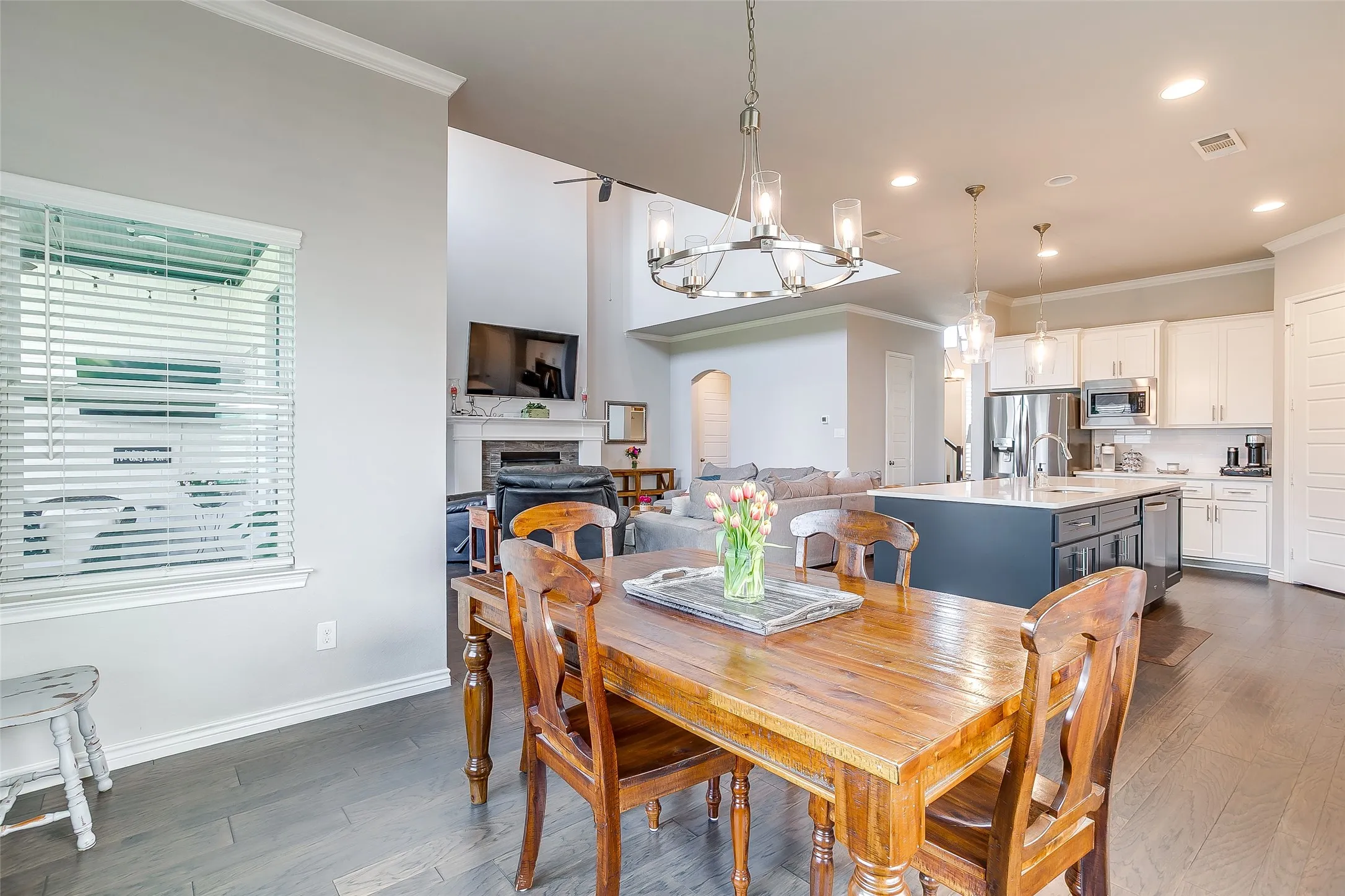 Open-concept living area featuring wood-finish flooring, a dining space with chandelier lighting, a kitchen with a central island and white cabinetry, a fireplace with a mantel, and a two-story ceiling in the background
