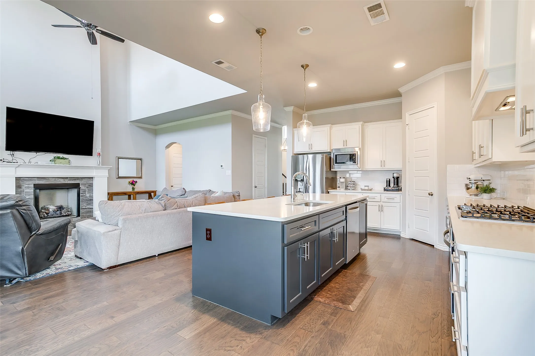 Open-concept kitchen and living area featuring wood-finish flooring, a kitchen island with pendant lighting, stainless steel appliances, and a stacked stone fireplace with a white mantel